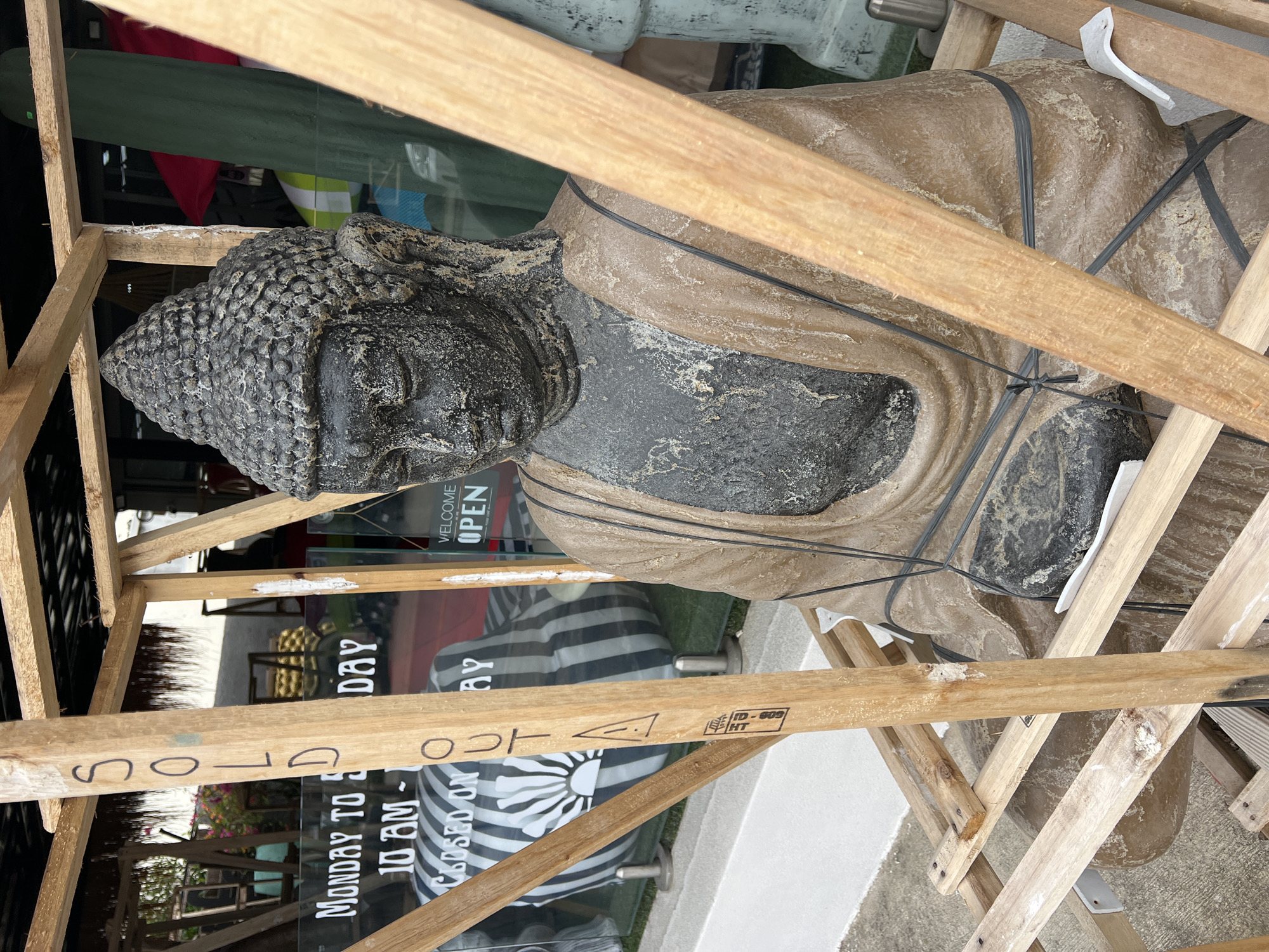A Buddha statue is secured inside a wooden crate in front of a storefront, possibly a shop or gallery, in Baan Bo Phut, Thailand. The statue, in shades of gray and tan, is wrapped with black straps. The unpainted wooden crate is crudely constructed. Through the store's glass windows, a sign partially reads "MONDAY TO..." and "WELCOME OPEN." On the wooden crate, the words "SOLD" and "OUT" are visible, along with "ID-609 HT." A glimpse of the storefront interior shows a striped cushion and possibly other merchandise. The surroundings are urban, with a paved surface visible.