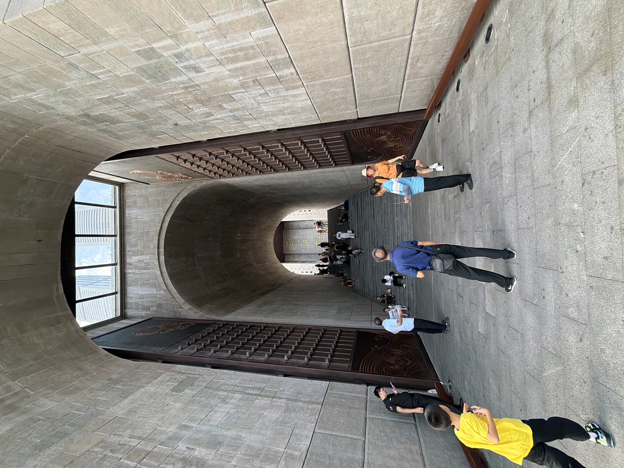 The image depicts the entrance to a large structure, possibly a museum or historical site, in Nan'an District, China. The architecture is imposing, with massive stone walls and an arched passageway leading to a wide staircase filled with people. The stonework is rough-hewn, suggesting an ancient or historically inspired design.
In the foreground, a young boy in a bright yellow t-shirt and black pants is captured in mid-motion, looking towards the left side of the frame. To his right, a man in a black uniform, possibly a security guard, is looking down at a tablet device. Further back, a man in a blue shirt holds a pamphlet, and another man in a blue and white striped shirt stands with his back to the camera, holding a similar item. On the right side of the frame, a woman in an orange top and black shorts is walking up the stairs, and a man in a blue shirt walks beside her.
The stairs are lined with people, some sitting and others walking up. The overall atmosphere appears to be one of a public gathering or tourist attraction. The structure is framed by tall, ornate doors on either side of the entrance. The sky is visible through a large opening at the top of the arch, revealing modern skyscrapers in the distance, indicating the juxtaposition of historical architecture with the contemporary urban environment. The lighting suggests it is daytime, possibly late afternoon, with shadows casting across the stone steps.