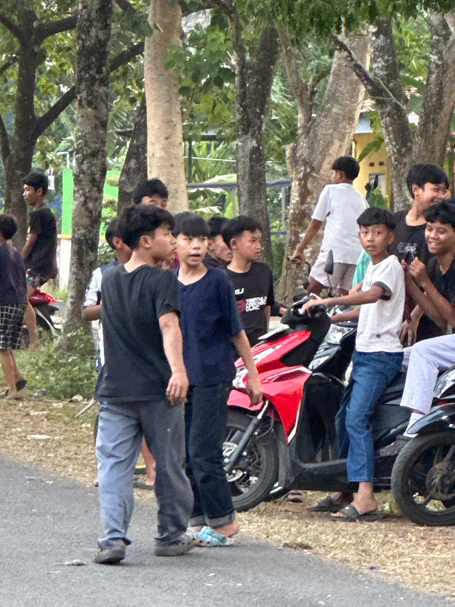 In Saruni, Indonesia, a group of young men and boys are gathered outdoors on a paved road adjacent to a grassy area with trees. Several individuals are standing around and on motorcycles. One boy in the foreground is sitting on a red motorcycle, wearing a white t-shirt and blue jeans. To his left, another boy in a black t-shirt and grey pants is standing, looking to the side. Behind them, other young men are also visible, some standing and some partially obscured by the motorcycles. The environment suggests a casual, informal gathering.