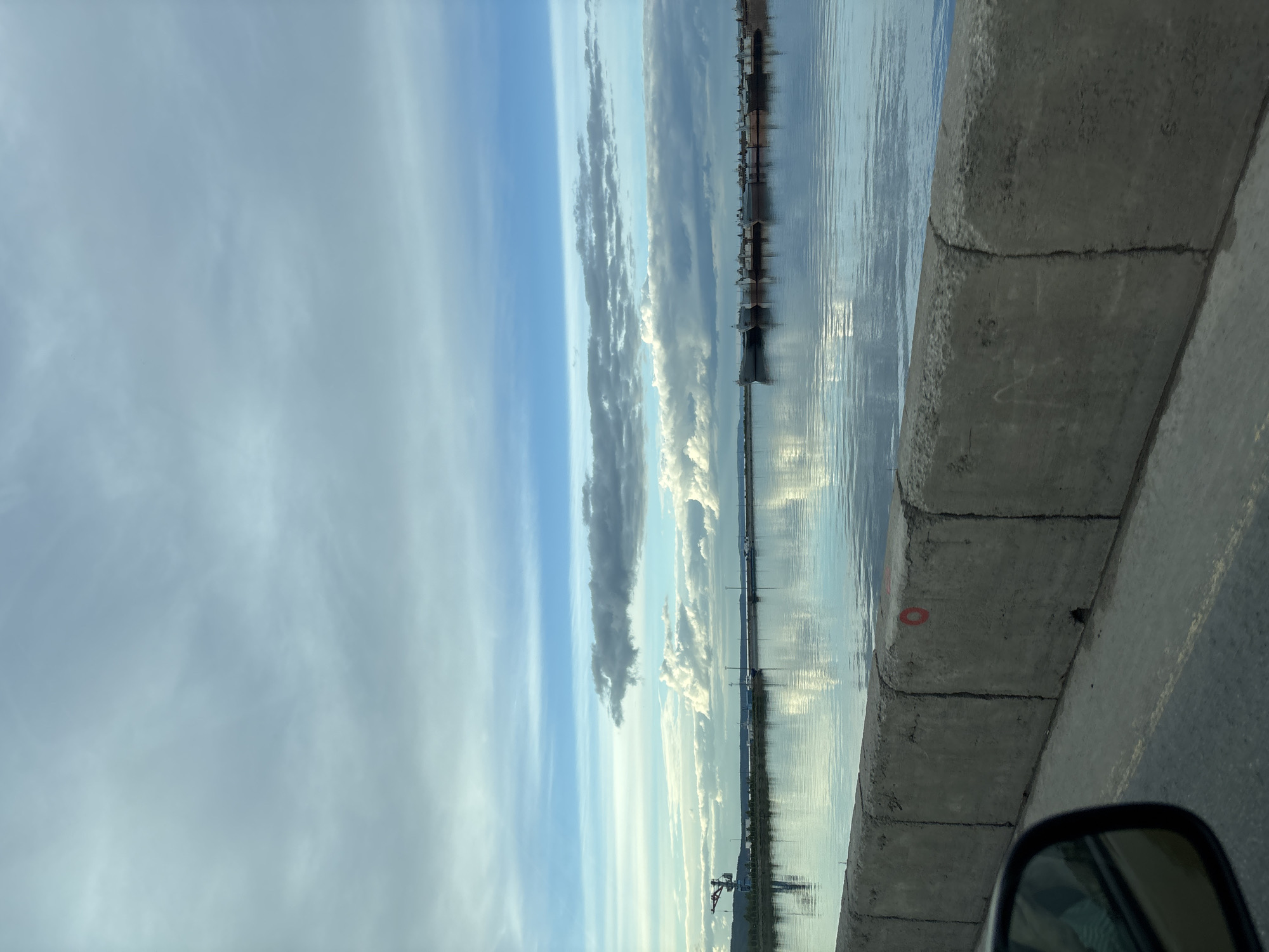 The image shows a waterfront view in what is presumed to be Chaikovsky, Russia. A concrete barrier runs along the right side of the frame, likely separating a road from the water's edge. The water reflects the sky, which is filled with elongated clouds. In the distance, there appears to be some kind of industrial infrastructure along the waterfront. A rearview mirror is partially visible in the lower right corner, suggesting the photograph was taken from inside a vehicle. The scene is dominated by gray and blue tones.