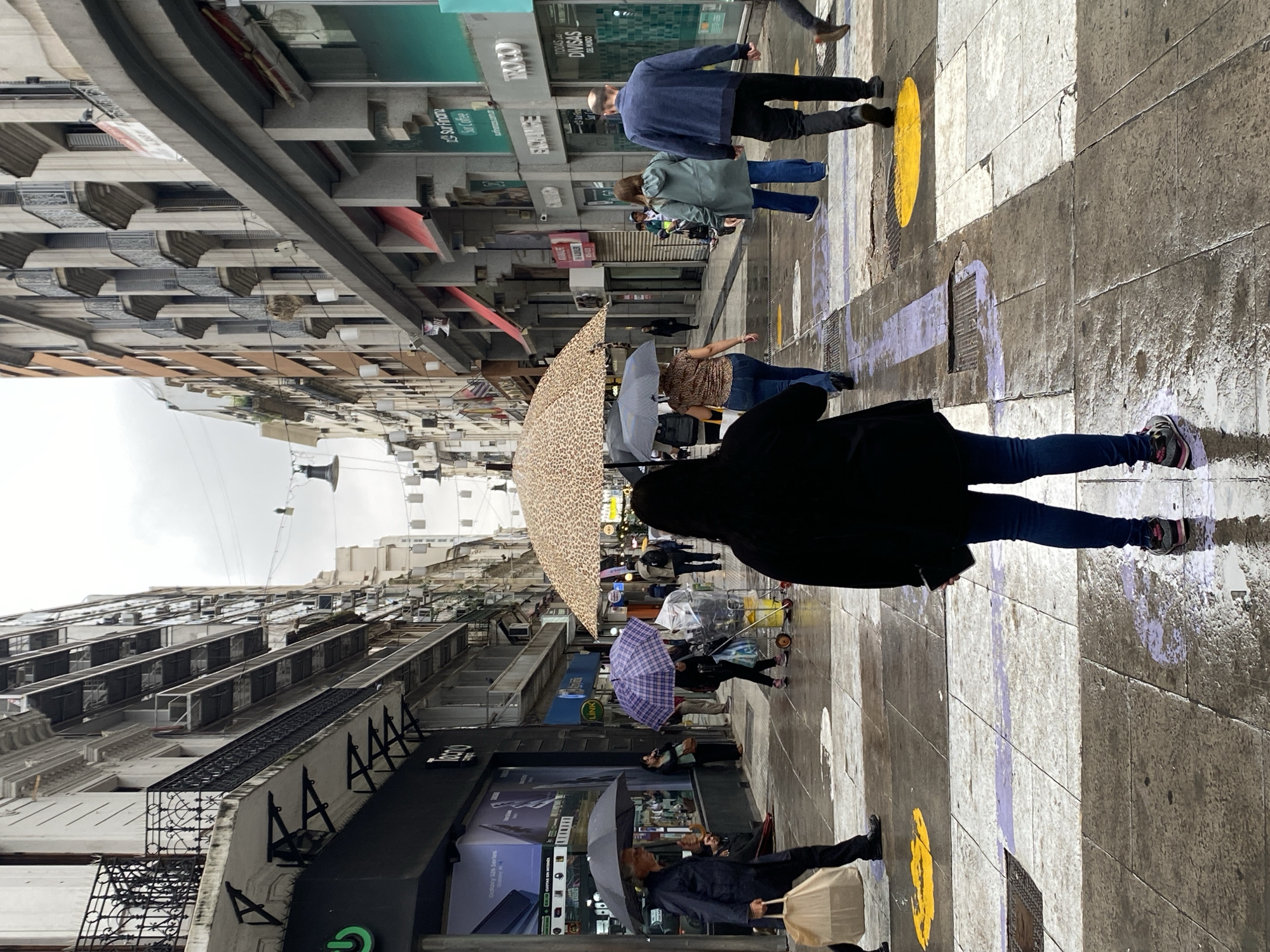 The image shows a street scene in Buenos Aires, Argentina on a wet day. Pedestrians are walking on a zebra crossing, several with umbrellas. The street is lined with buildings, including shops, and the reflections in the wet pavement are visible. Graffiti or painted markings are on the road. People carry umbrellas of various designs. A shop sign reads "ROCO TODAS LAS DIVISAS DEL MUNDO", and there is a shop with electronics, probably a cellular phone store. The street appears to be a busy commercial area.