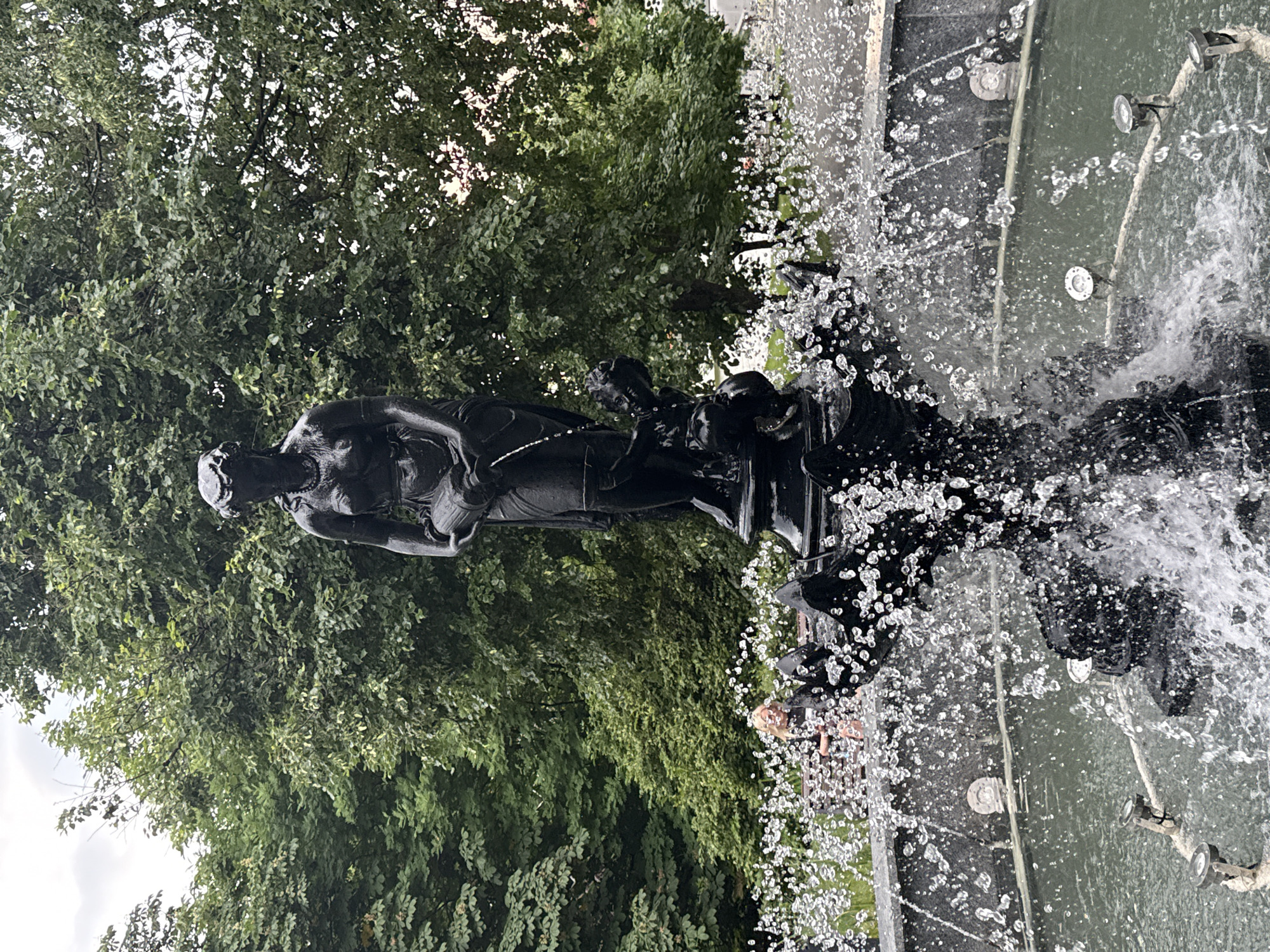 The image shows a fountain in a park with lush green trees in the background. The fountain features a dark bronze sculpture of a woman holding a jug and a smaller figure at her feet. Water sprays upwards and outwards from the fountain, creating a dynamic visual effect. In the middle ground, a woman with blonde hair is seated on a bench, looking towards the fountain. The overall scene appears to be a serene outdoor setting, likely a public park or garden. Based on stylistic elements and common public art found in Moscow, this fountain is likely located in the Neskuchny Garden or Gorky Park.