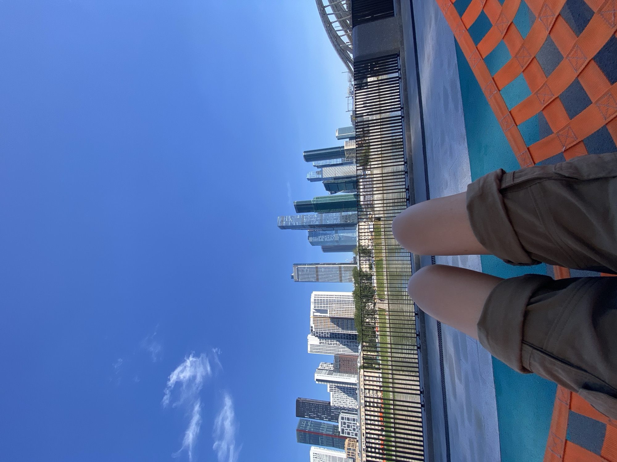A first-person perspective shot shows a person's legs from the knees down, wearing khaki shorts, resting on an orange net hammock. The background features a bright blue sky with a few wispy clouds, a body of water, and a cityscape dominated by modern skyscrapers. The skyline includes prominent buildings like the Federation Tower, Mercury City Tower, and Naberezhnaya Tower, identifying the location as Moscow City, Russia. A black metal fence separates the hammock area from a promenade, beyond which lies a river and the city's financial district. The overall impression is of a relaxed outdoor scene in a bustling urban environment.
