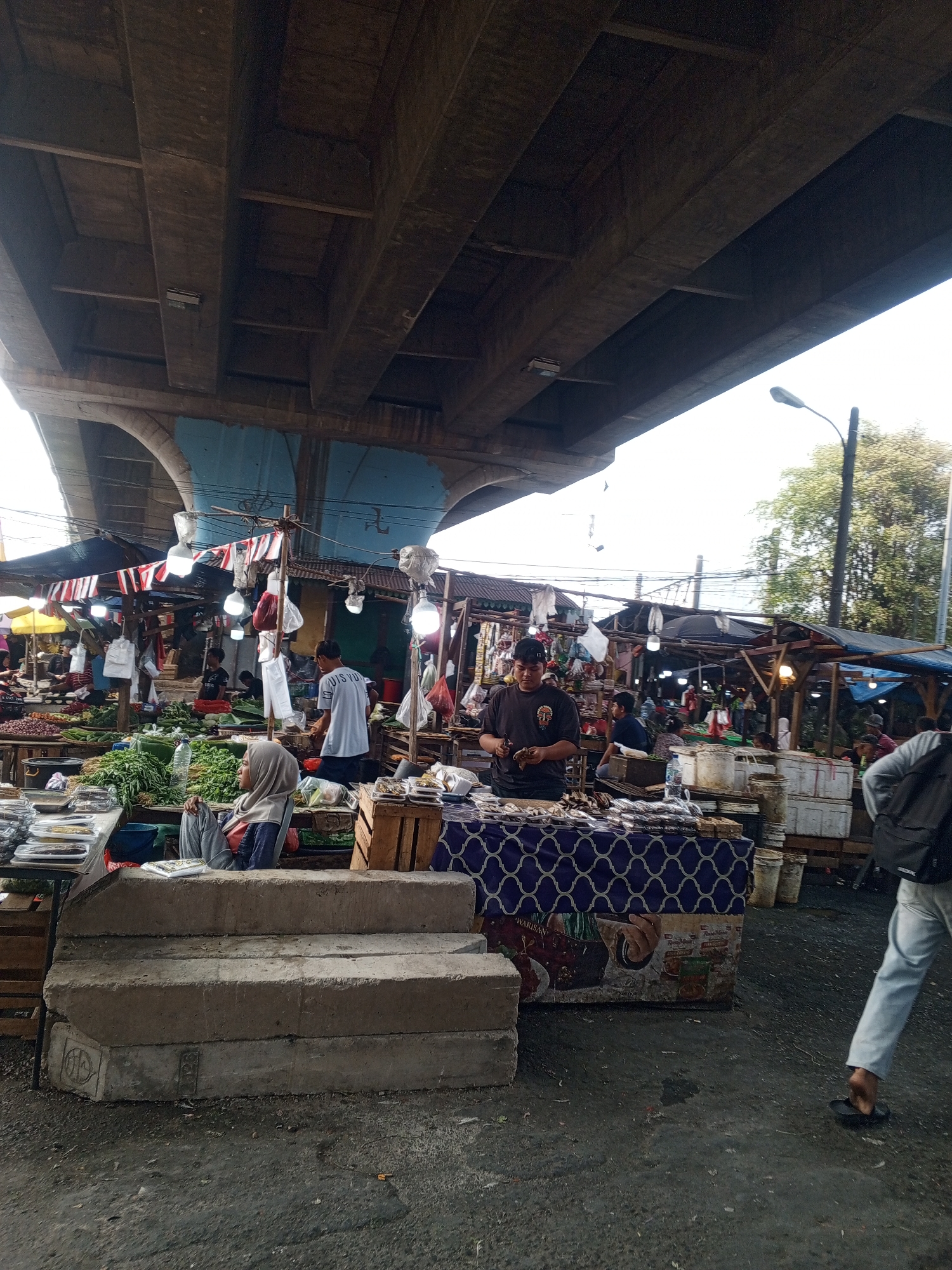 The image captures a bustling market scene under a large overpass in Depok, Indonesia. The overpass, constructed of concrete beams, dominates the upper portion of the frame. Below, a vibrant market unfolds with stalls displaying a variety of produce, including an abundance of green vegetables. Several people are present, engaged in various activities. In the foreground, a woman wearing a grey hijab and matching trousers sits on concrete blocks, appearing to observe the market. To her right, a man in a dark t-shirt is busy at a stall, possibly preparing or selling goods. Another man, wearing a light blue t-shirt with the words "LOUIS VUITTON" is visible further back, interacting with the market environment. Various market stalls with tarpaulin coverings are set up, adorned with decorative flags and illuminated by hanging lights. Wooden crates and plastic containers are used to display and store goods. The ground is a rough, asphalt-like surface, typical of outdoor markets. A street lamp is visible on the right side of the image, suggesting it is a public thoroughfare. The overall atmosphere is one of lively commerce within an urban setting.