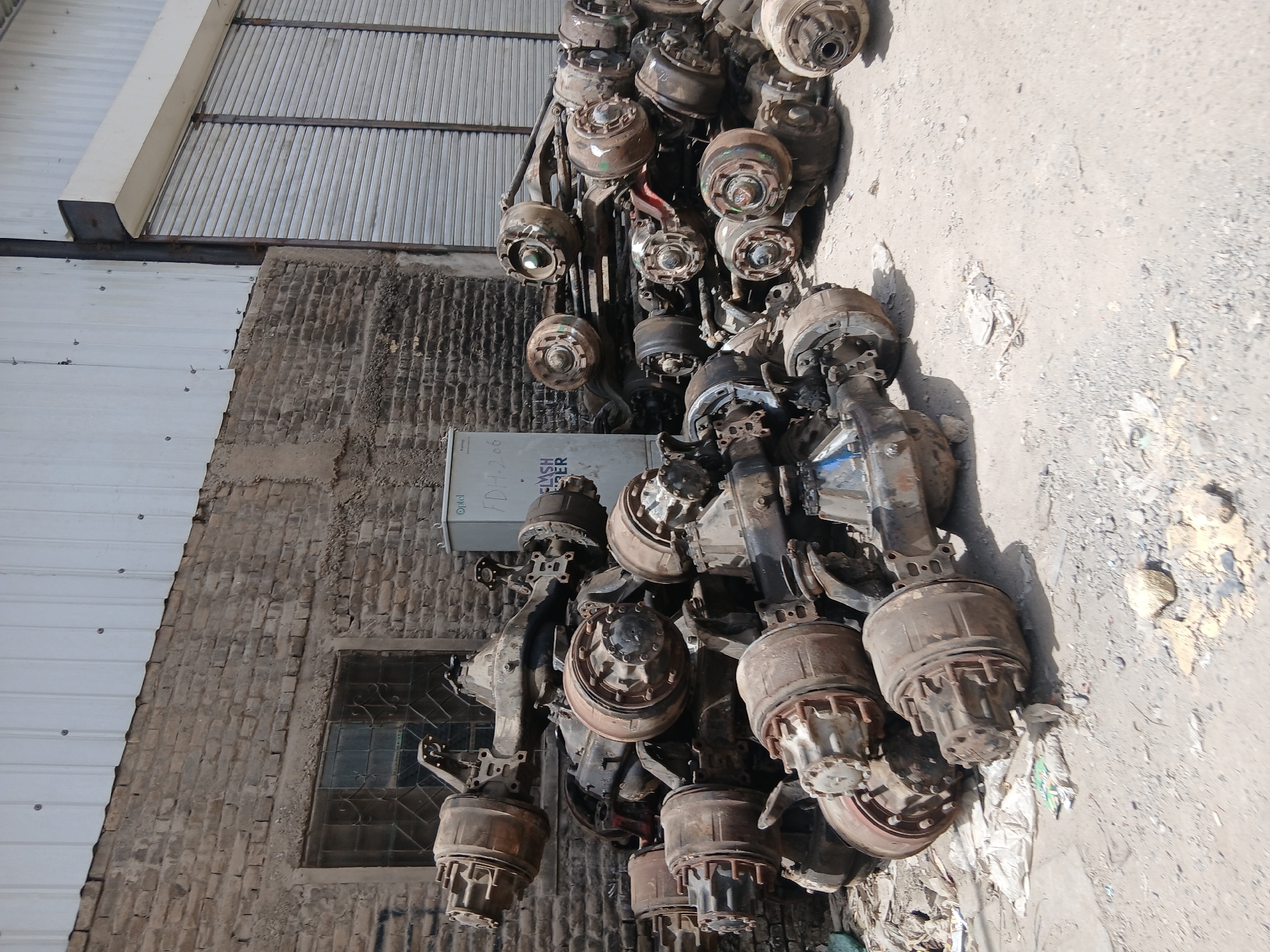 The image depicts a pile of used truck axles and brake assemblies. They are located on an uneven, gray concrete or asphalt surface. In the background, there is a building with a corrugated metal roof and a brick wall featuring a window with ornate metal bars. The building also has an electrical box mounted on the wall. Debris and discarded materials are scattered on the ground. Given the location, Quetta City Tehsil, Pakistan, and the presence of truck parts, it is likely an automotive scrap yard or a business dealing with vehicle parts. There are no people visible in the image.