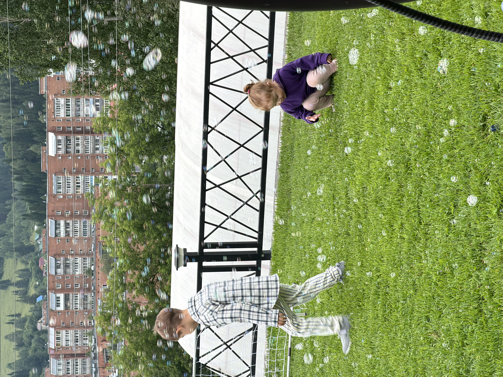 Two children are playing in a grassy area with many bubbles. A boy in a plaid shirt and striped pants is walking from left to right. A younger girl in a purple hoodie and light-colored pants is squatting and looking at the ground. In the background, there is a black metal fence with a white fabric structure behind it. Beyond the fence, there are trees and a row of brick apartment buildings, characteristic of the urban landscape of Tashtagol, Russia. The sky is overcast.