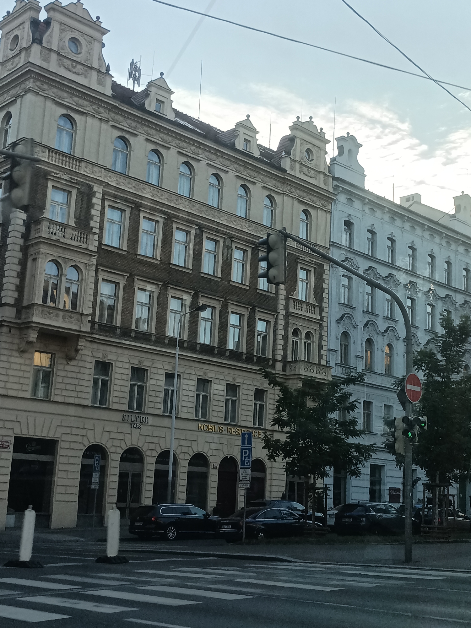 The image shows a street scene in Prague, Czechia, with ornate European-style buildings lining the street. The buildings feature multiple stories with distinct architectural details, including arched windows, balconies, and decorative facades. On the left, a darker brick building is juxtaposed with a lighter, possibly cream-colored building to its right. Further down the street, a pale blue building with intricate detailing is visible. Traffic lights are present, with one showing a green light, indicating that traffic can proceed. Several cars are parked or driving on the street, and a pedestrian crossing is marked on the road. Trees are planted along the sidewalk, and overhead wires suggest the presence of streetcar or trolley lines. The sky is overcast, suggesting a cloudy day. Signs are visible on the buildings, including "SILVER TAPS" and "NOBILIS RESIDENCE". A red and white circular sign with a horizontal bar is mounted on a traffic light pole.