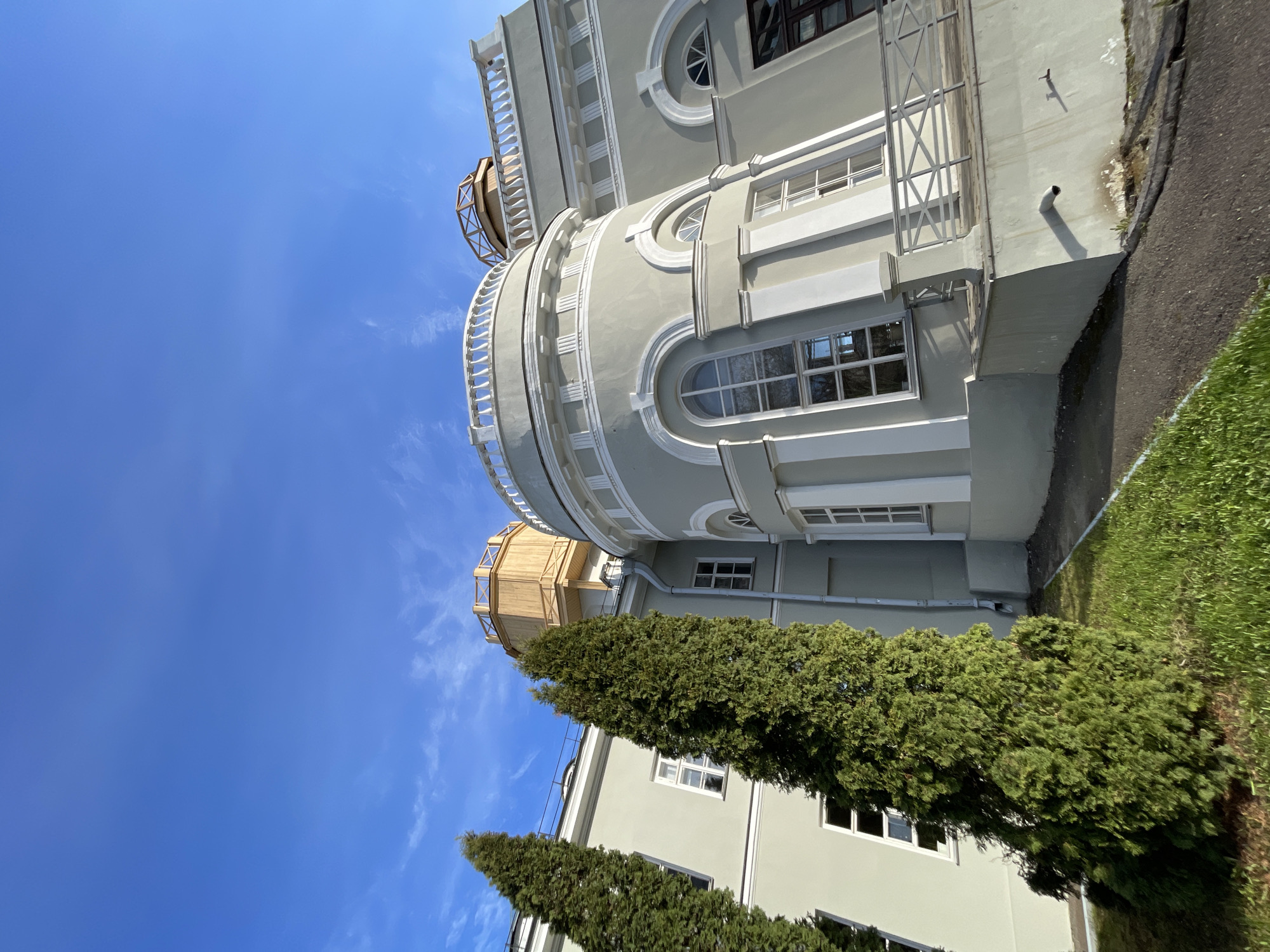The media file presents a large, architecturally ornate building under a clear, bright blue sky with faint wispy clouds. The building is painted in a light gray or pale green color, adorned with prominent white decorative elements such as cornices, pilasters, and balustrades. It features distinct rounded sections with arched windows, alongside numerous rectangular windows.
On the roof of the rounded building sections, there are structures resembling observatory domes. One is clearly visible, constructed from light-brown wood or wood-like material with a slatted or lattice design.
In the foreground and midground, several tall, dark green evergreen trees (likely cypresses or similar conifers) partially obscure the lower parts of the building. To the right, a strip of vibrant green grass runs alongside a dark paved path or driveway. Some external piping is visible on the building's facade.
The image depicts a static scene on a sunny day, evidenced by the bright light and sharp shadows cast by the architecture and trees. No people or activities are visible. The setting conveys a sense of calm and historical significance, potentially indicating a scientific or academic institution, aligning with the provided context of Kazan, Russia. No visible text is present in the image.