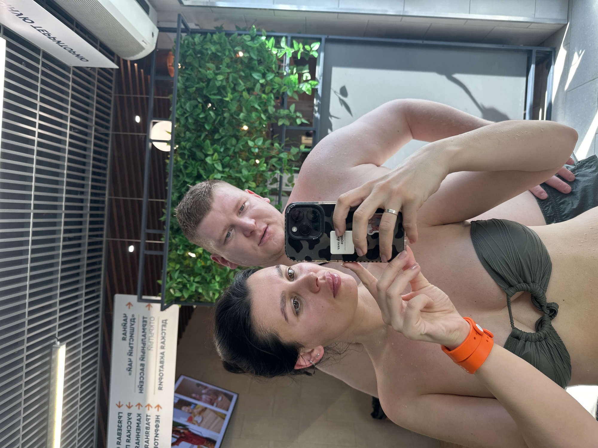 A man and a woman are taking a selfie in what appears to be a sauna or spa area. The woman is holding the phone, wearing a green bikini top and an orange wristband. The man is behind her, with water droplets on his skin. A green leafy plant is visible in the background, as well as a sign with text in Russian. The location is likely in Votkinsk, Russia, based on the presence of Russian text.