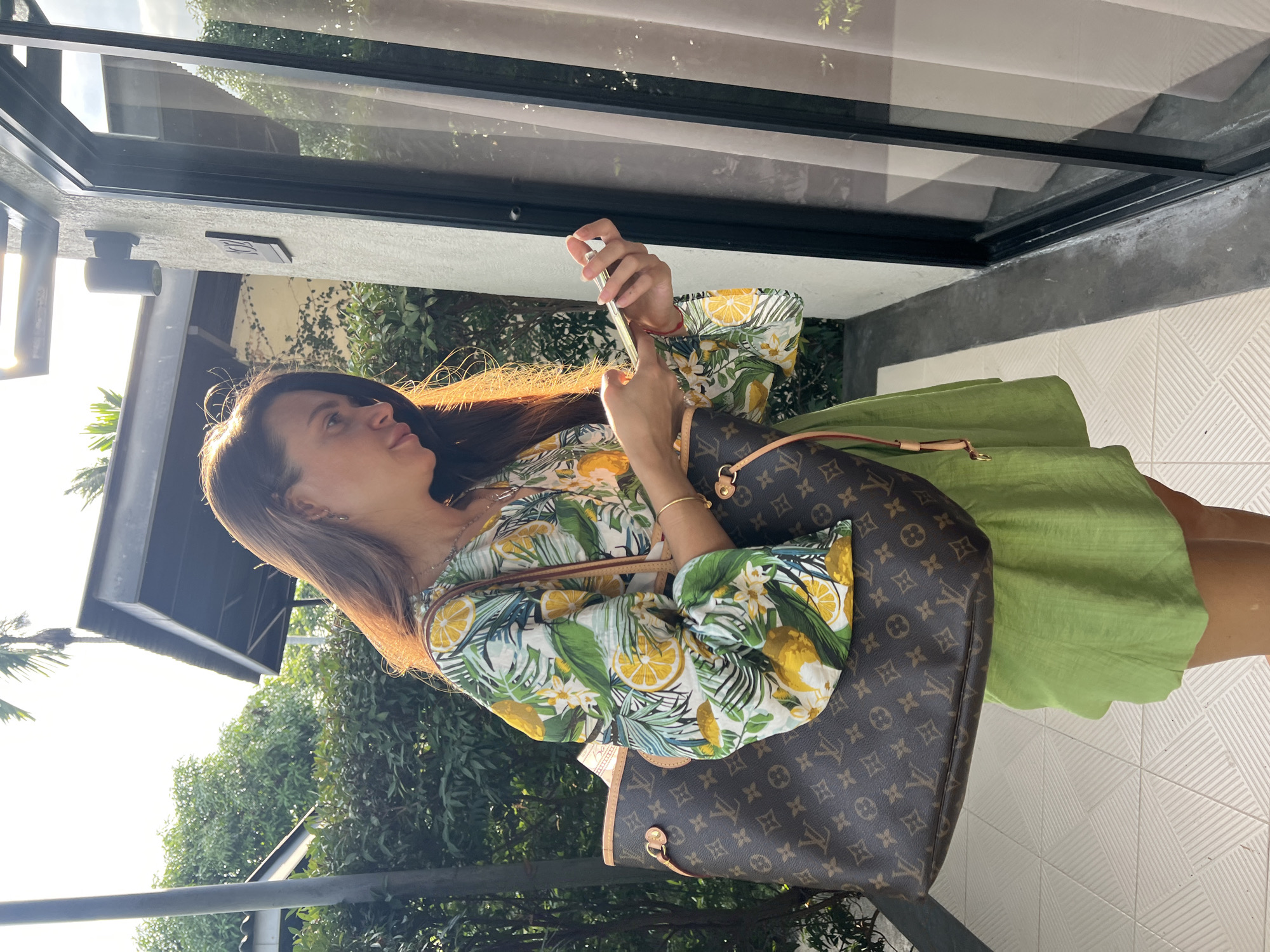 A woman stands on a tiled patio in บ้านบางรักษ์, Thailand, holding a phone and looking at it. She is wearing a light green dress and a white shirt with yellow lemons. She has a Louis Vuitton handbag slung over her shoulder. Behind her are tropical trees and a building with large glass windows. The scene is well-lit.
