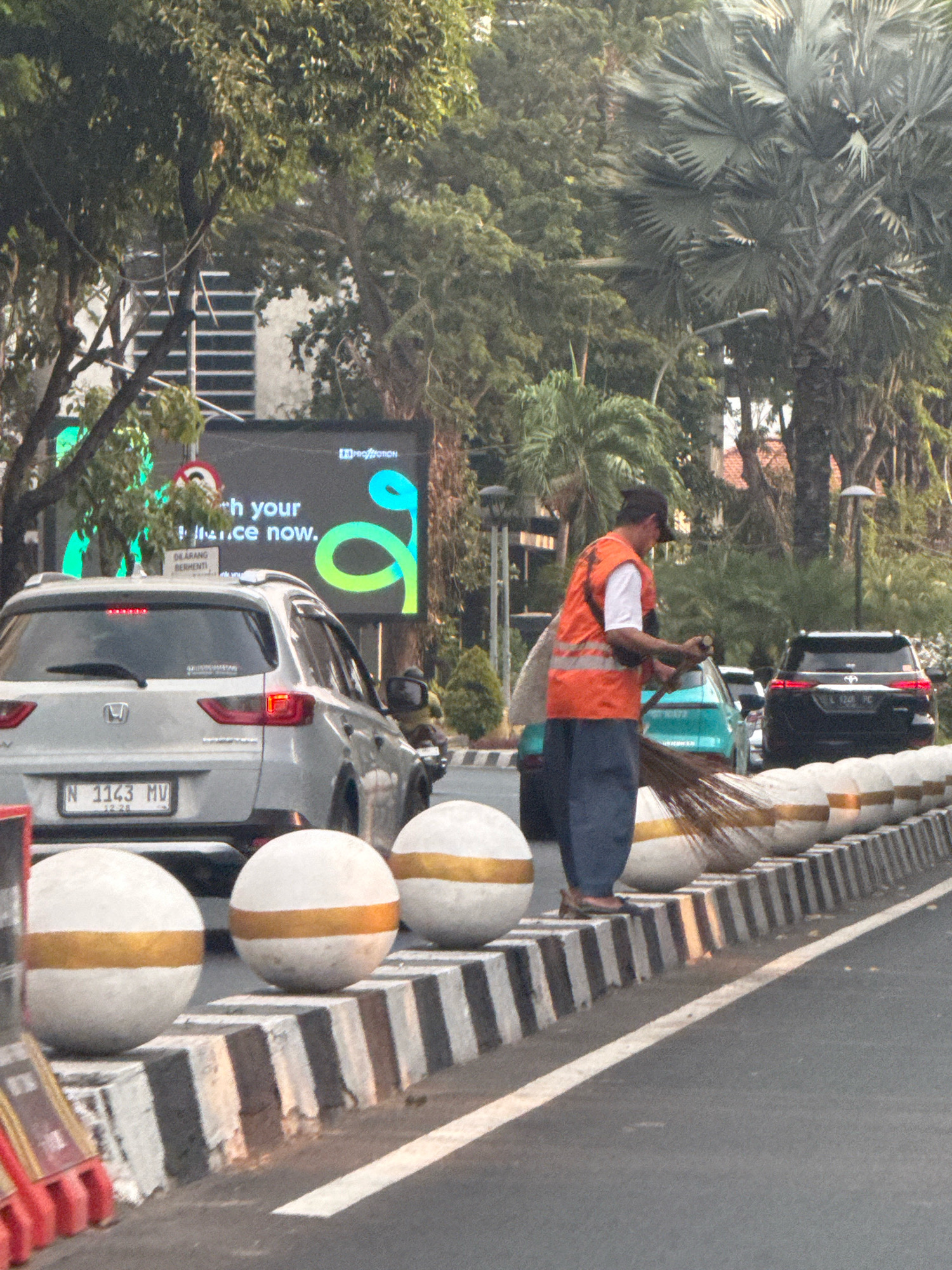 A street sweeper in an orange vest and dark pants is sweeping the side of a road in Surabaya, Indonesia. He is holding a broom and appears to be focused on his task. In the background, there are several cars, including a silver Honda SUV and a black Toyota SUV. There is also a large digital billboard displaying an advertisement. The street is lined with decorative concrete barriers topped with white spherical bollards, each with a gold band. Trees and foliage are visible throughout the scene, suggesting a tropical or subtropical climate. The sky is overcast, and the overall lighting suggests it is daytime. The image captures a mundane urban moment, highlighting the presence of essential services within the city's infrastructure.
