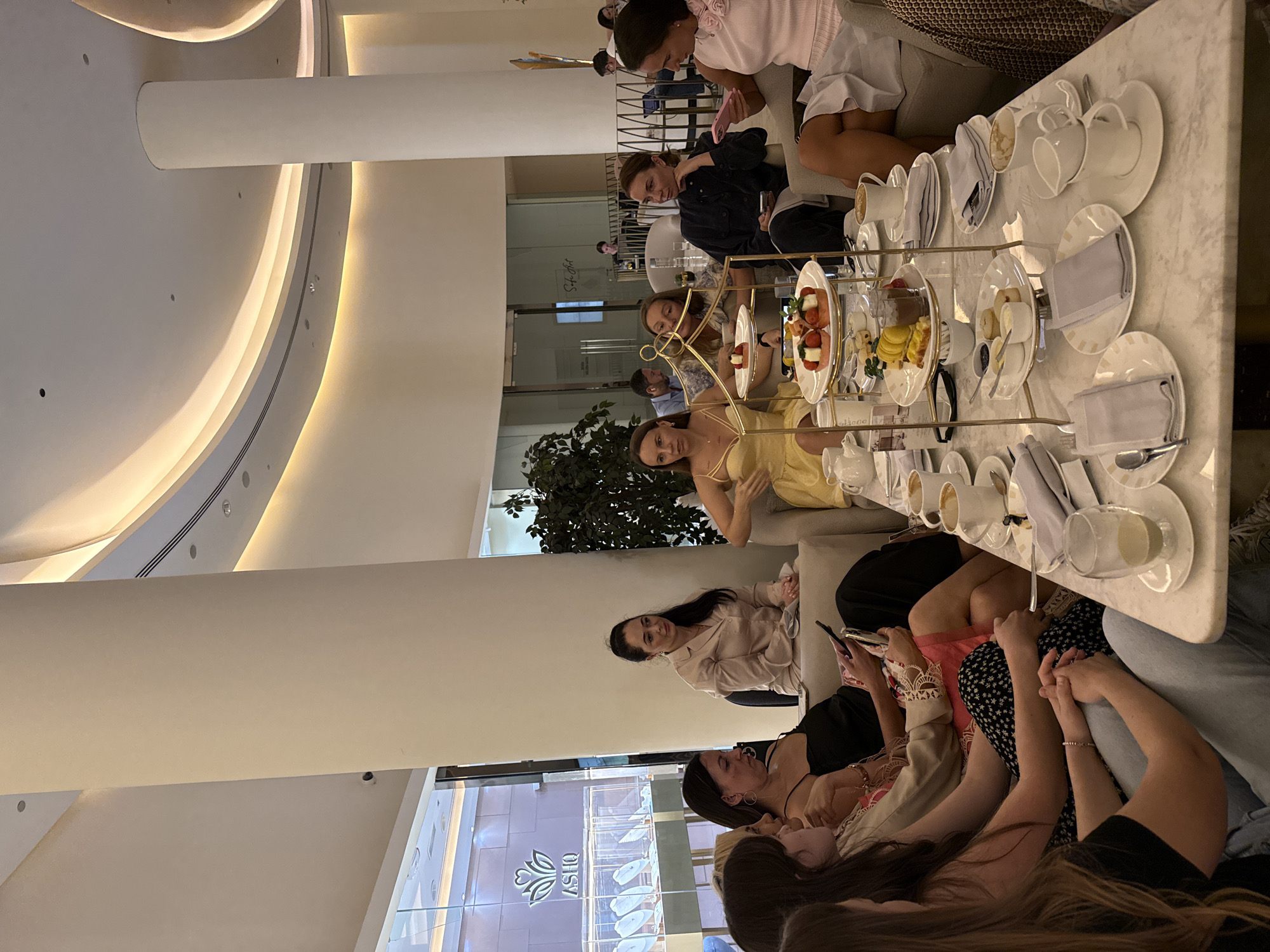 This image depicts a social gathering, likely an afternoon tea or coffee session, in a contemporary and upscale indoor setting. The scene is rotated 90 degrees counter-clockwise.
The setting features a modern, high-ceilinged interior with prominent architectural elements including large, smooth, curved white ceilings and substantial cylindrical white pillars. Warm, indirect lighting emanates from recessed fixtures and coves along the ceiling and architectural contours, creating a sophisticated and inviting atmosphere. The spacious design suggests a cafe or lounge within a larger complex, possibly a luxury mall or hotel in Dubai.
A group of women, appearing to be young to middle-aged adults, are seated around a long, light-colored marble-patterned table and on comfortable sofas. They are engaged in various activities: some are conversing, others are looking at their mobile phones. One woman in a light-colored blouse looks directly towards the camera, while another in a yellow dress looks upwards. A few men are subtly visible in the background.
The table is meticulously set with an assortment of delicacies typical of afternoon tea. Multiple tiered serving stands display various small treats including mini sandwiches, fresh fruit (such as strawberries and berries), macarons, small cakes, and scones, accompanied by jam and cream. Numerous white ceramic cups and saucers, some containing coffee or tea, are arranged on the table alongside plates with neatly folded napkins and cutlery.
Notable details include the elegant and minimalist design of the venue, with its clean lines and bright surfaces. The visible text "ASHQ" appears on a glass display cabinet in the background, suggesting a nearby luxury retail store, which aligns with the upscale nature of the setting in Dubai. The lighting indicates an indoor gathering, possibly during the daytime or evening, emphasizing comfort and leisure.