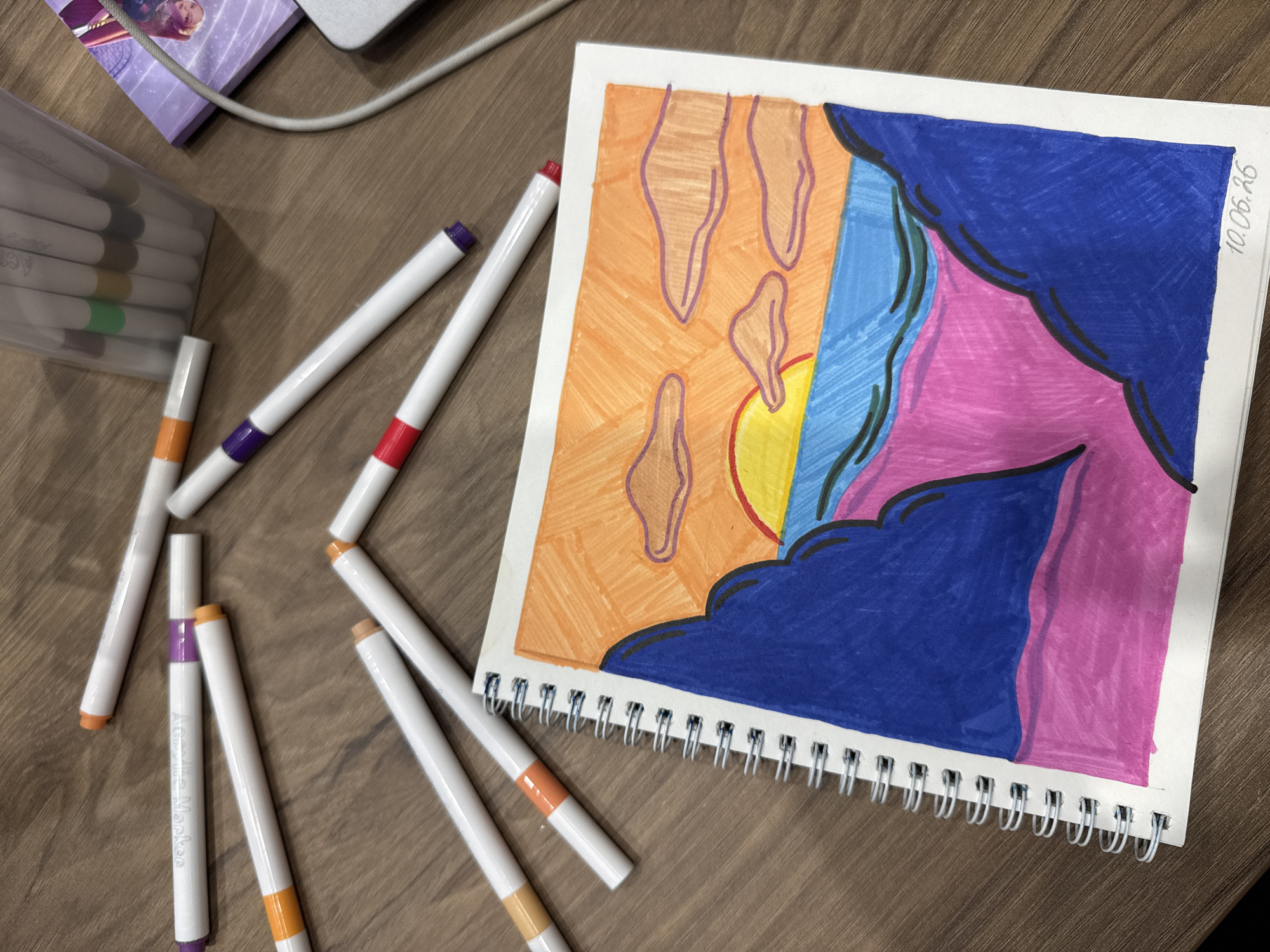 The image depicts a close-up overhead view of a spiral-bound sketchbook containing a colorful drawing of a sunset over the ocean. The drawing features orange and yellow hues for the sky and sun, blue for the water, and large dark blue shapes representing mountains or landmasses in the foreground, with a pink area below. Scattered around the sketchbook are several white markers with colored tips, including purple, orange, and red. In the background, a clear plastic case holding more markers and a portion of a purple notebook with a character illustration are visible. A gray computer cable is also present. The entire scene is set on a brown wooden surface. The image does not contain any identifiable landmarks or elements that would indicate a specific location within the city of Murom, Russia.