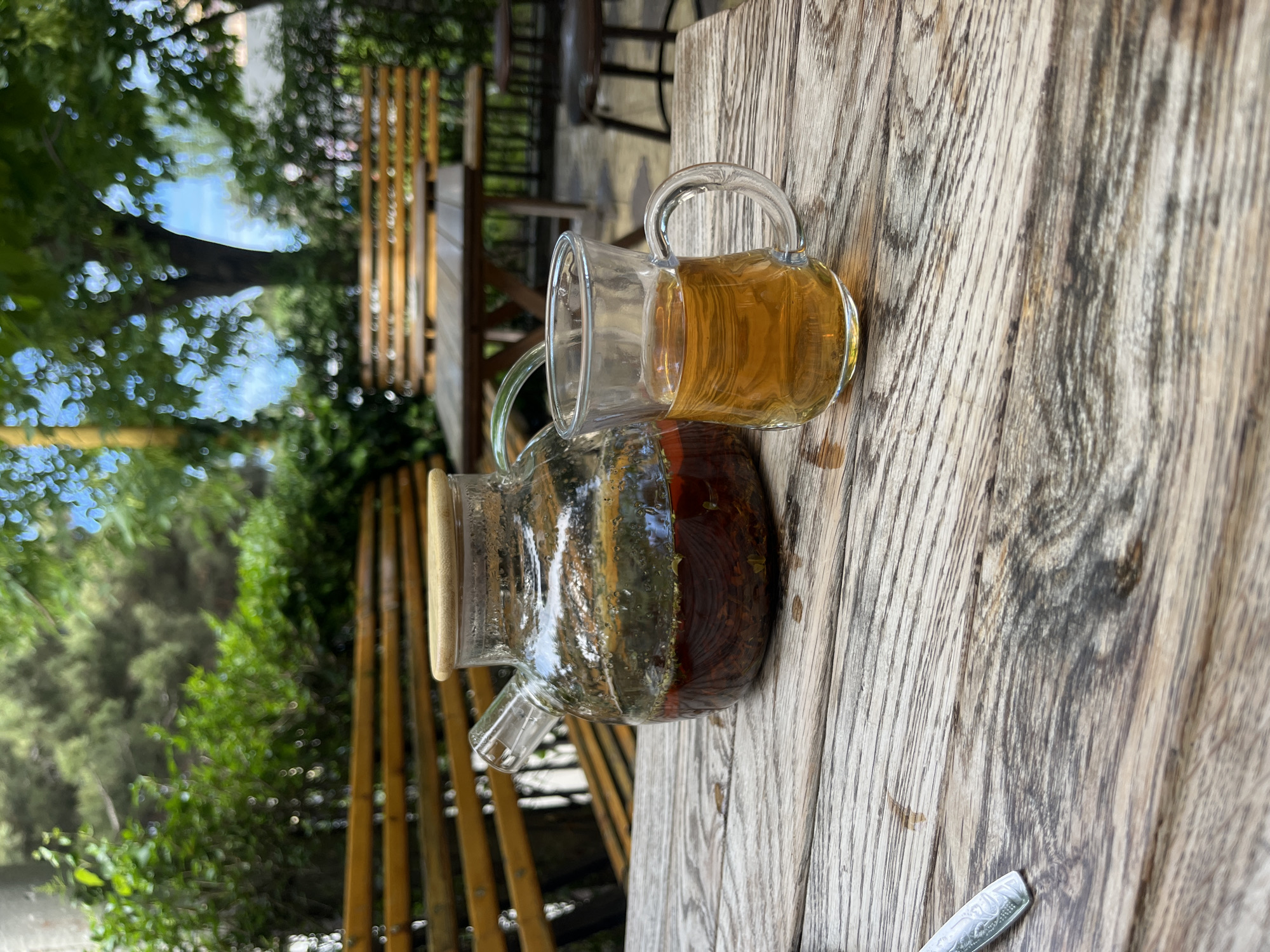 On a weathered wooden table in Kaspiysk, Russia, sits a glass teapot partially filled with dark tea, alongside a matching glass mug holding a lighter-colored tea. Outside, a green canopy of trees and partial views of wooden benches and fencing suggest an outdoor cafe or patio setting. The scene is captured from a slightly elevated perspective, showing the table and objects in clear focus against the blurred background.