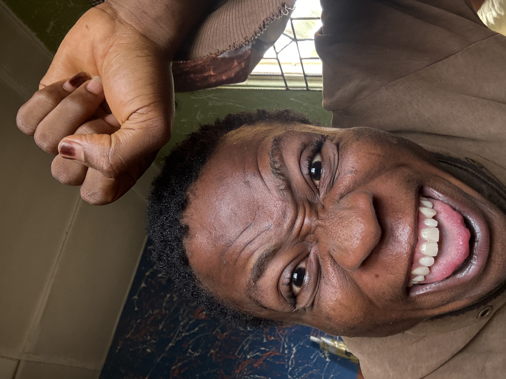 A close-up, distorted view of a person's face with an exaggerated expression of happiness or excitement. The person is dark-skinned with short, black hair. Their eyes are wide, and wrinkles are visible on their forehead and around their eyes. The person's mouth is wide open, revealing their teeth and tongue. The person is wearing a brown hooded sweatshirt. A hand with short, manicured nails is visible in the upper left corner of the frame. The background is blurry, with glimpses of a window and a wall. The location is Unknown, Unknown.