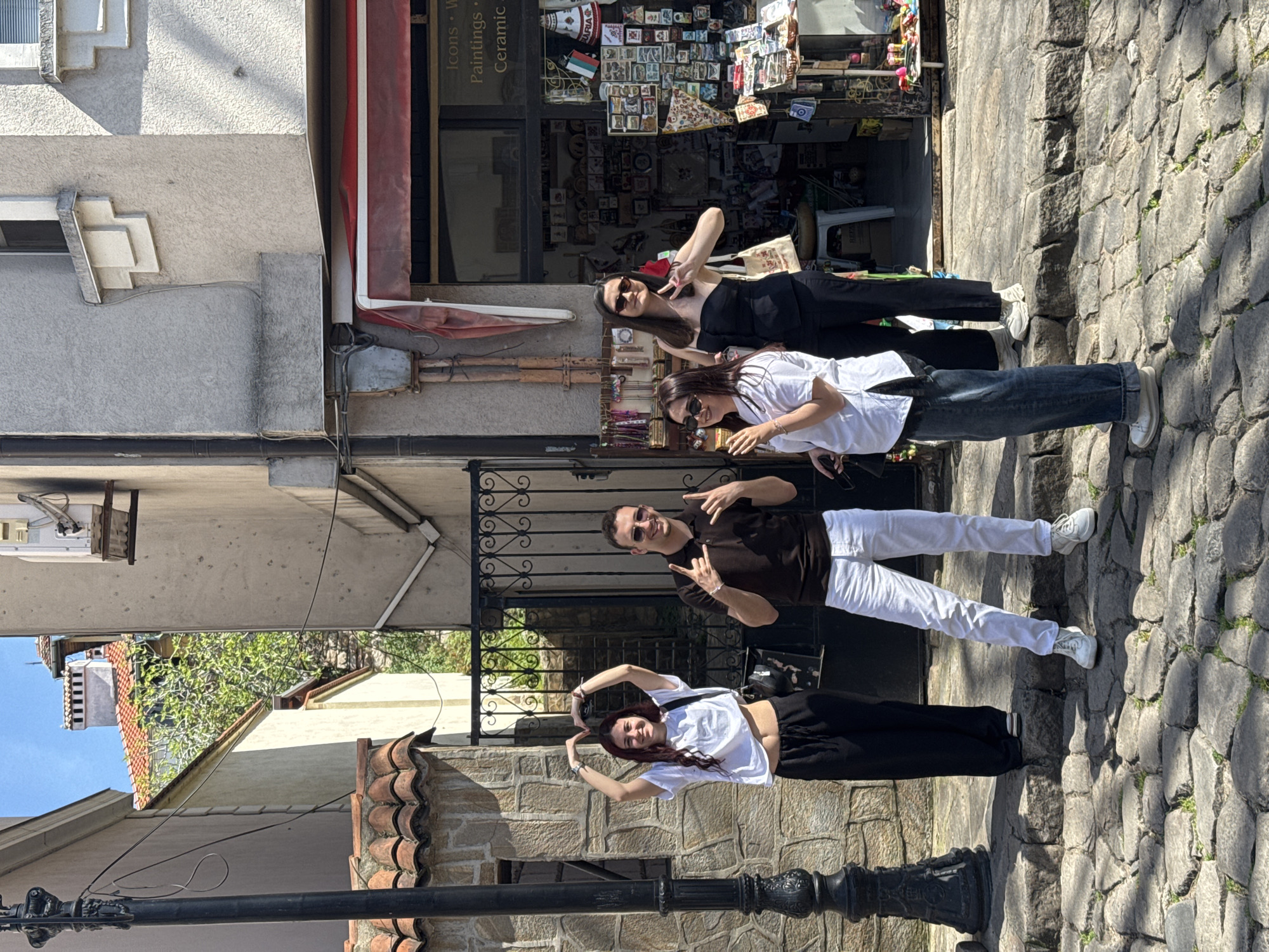 Four people are standing on a cobblestone street in Plovdiv, Bulgaria, posing for a photo.
From top to bottom:
1. A woman in sunglasses and a black outfit is throwing up a peace sign.
2. A woman in sunglasses and a white shirt with blue jeans.
3. A man in sunglasses and a brown shirt with white pants is throwing up the rock on fingers sign.
4. A woman in a white shirt and black pants is making a heart shape with her hands.
Behind them is a shop selling Icons, Paintings, and Ceramic wares. Behind them are the roofs of buildings with red tiles, and a section of bright blue sky. In the bottom left of the image is a black lamp post. The street is made of cobblestones.