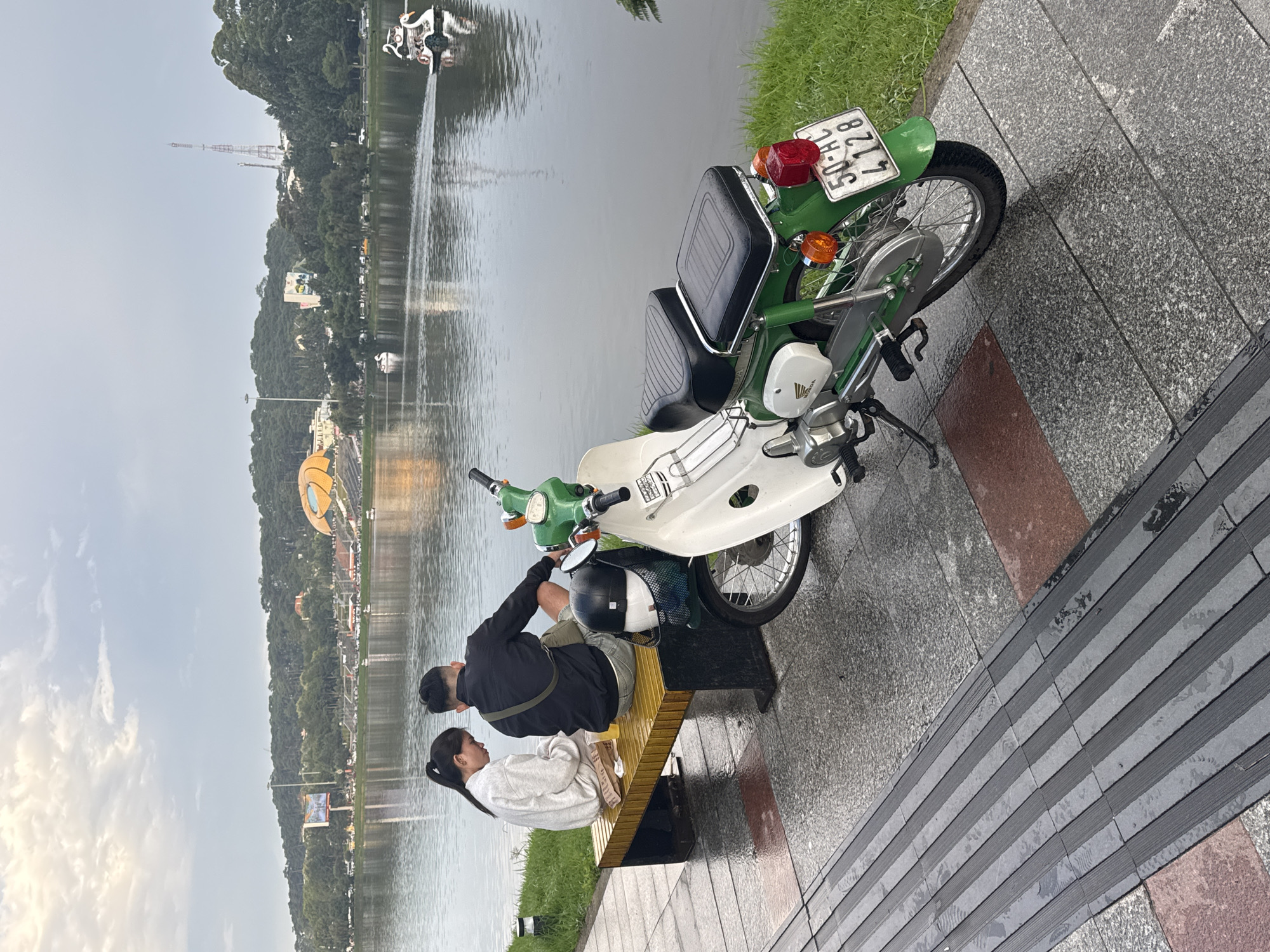In Da Lat, Vietnam, a couple sits on a yellow slatted bench beside Xuan Huong Lake. The woman, wearing a white hoodie, looks down at her hands while the man, in a black jacket and shorts, faces the lake. A green and white Honda motorcycle is parked next to the bench, with a helmet and bag on the seat. The lake is calm, reflecting the overcast sky and the lush green hills in the distance, dotted with buildings and a large, distinctive yellow flower-shaped structure. Swan-shaped pedal boats are visible on the water. The foreground shows a paved walkway with textured tiles, wet from recent rain.