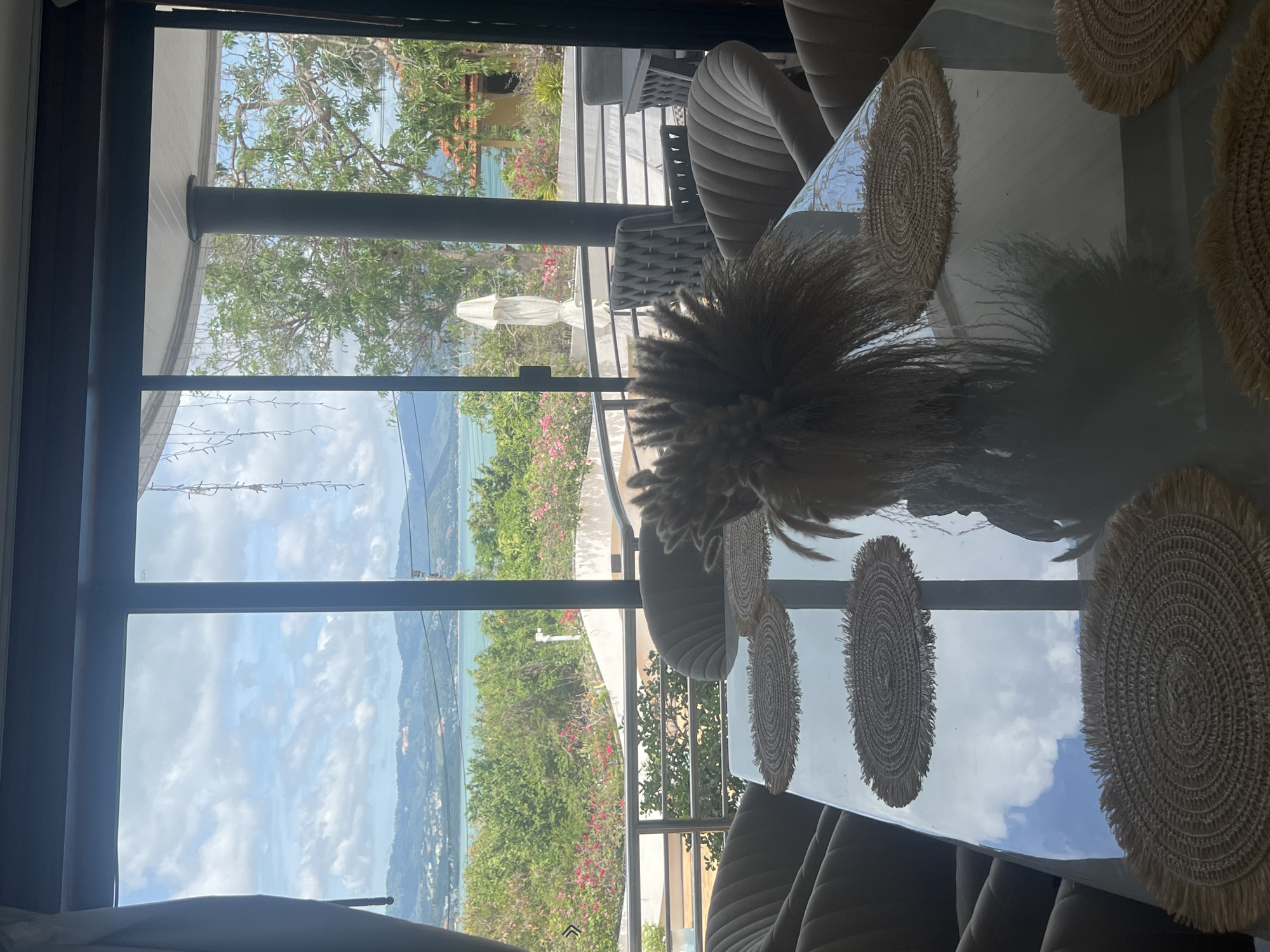 This image depicts a dining area with a large glass table situated in front of a wide window offering a view of the sea and a lush green landscape. The room features several plush dining chairs around the table. On the table, there are woven placemats and a decorative arrangement of dried grasses in a vase. The window reveals a scenic panorama of rolling hills, scattered buildings, dense foliage, and the turquoise ocean under a cloudy sky. The overall setting suggests a modern, relaxed dining space with a tropical ambiance, likely located in a coastal resort or private residence in Ko Samui District, Thailand, given the prominent view.