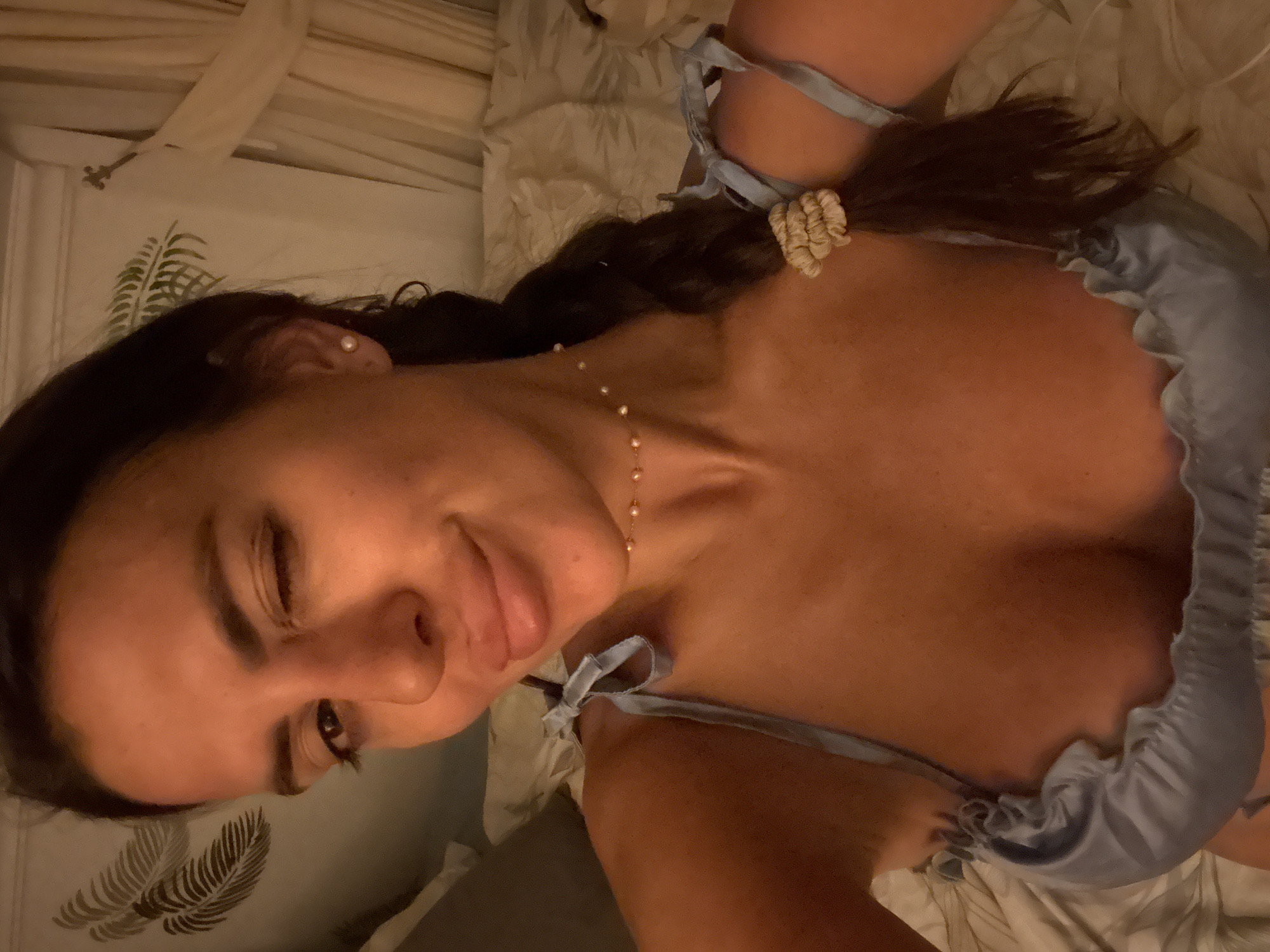 A woman is lying on a bed, winking at the camera. She has dark hair pulled back into a braid and is wearing a light blue silk camisole. A pearl necklace adorns her neck. The room is dimly lit, with sheer curtains in the background. The bed appears to be covered in a white comforter. There is a palm frond pattern on the wall behind her. The location is not specified as Fethiye, Turkey, or any other city.