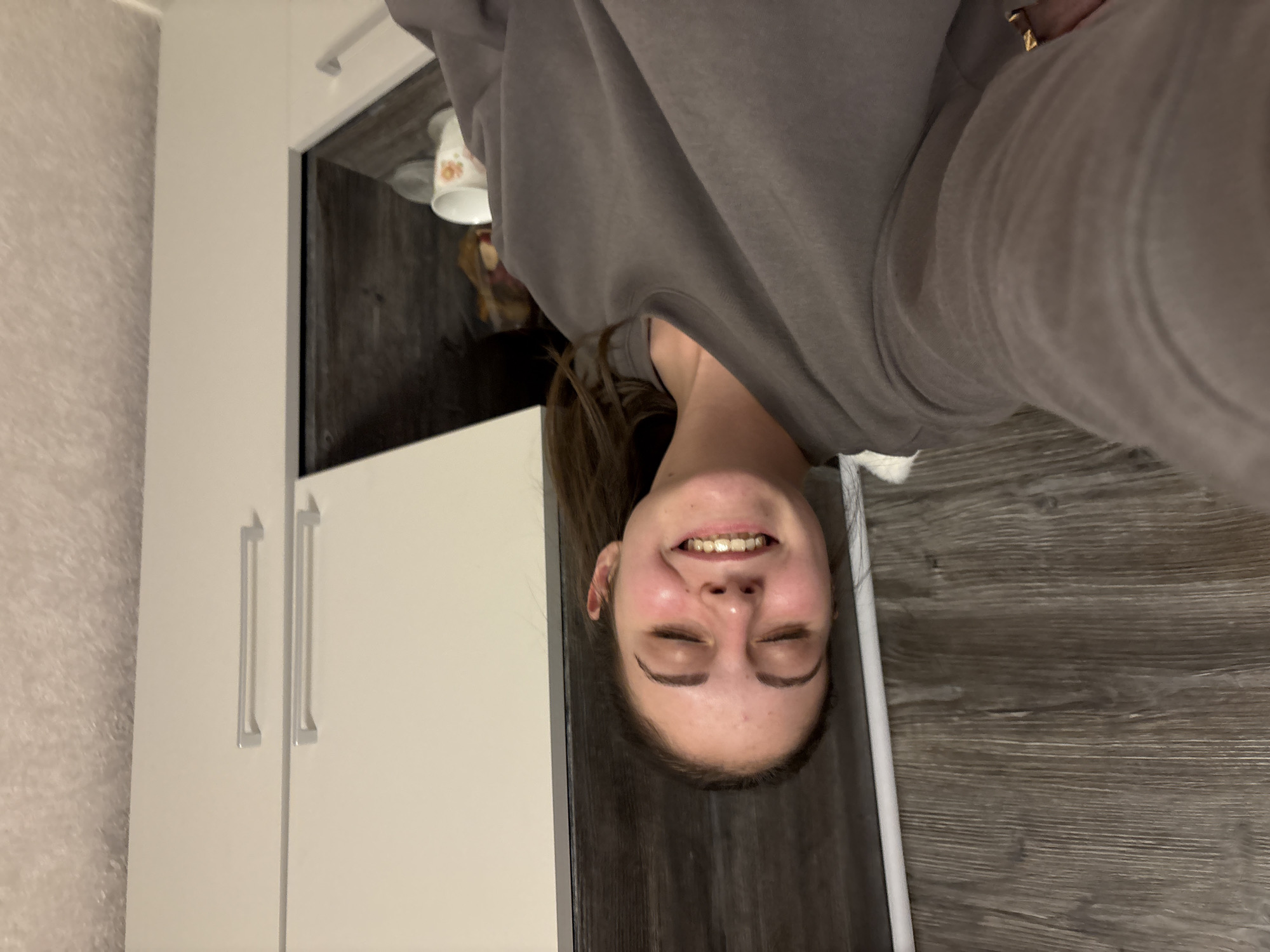 A young woman is captured in a joyful selfie, smiling broadly with her eyes mostly closed, conveying a sense of happiness or amusement. She has long dark hair and is wearing a plain grey, long-sleeved sweatshirt. A gold-colored ring is visible on her right hand.
The indoor setting features modern white cabinetry with sleek, elongated silver handles on the left side of the frame, adjacent to a light-colored, textured wall covering. A dark wood-effect surface, likely flooring or a countertop, occupies the right side and bottom of the background. On this surface, behind the woman, a white mug decorated with orange-brown patterns can be seen. The lighting in the room appears to be artificial and warm, contributing to a cozy ambiance. No discernible text is visible anywhere in the image. The scene is located in Bulgarovo, Bulgaria.