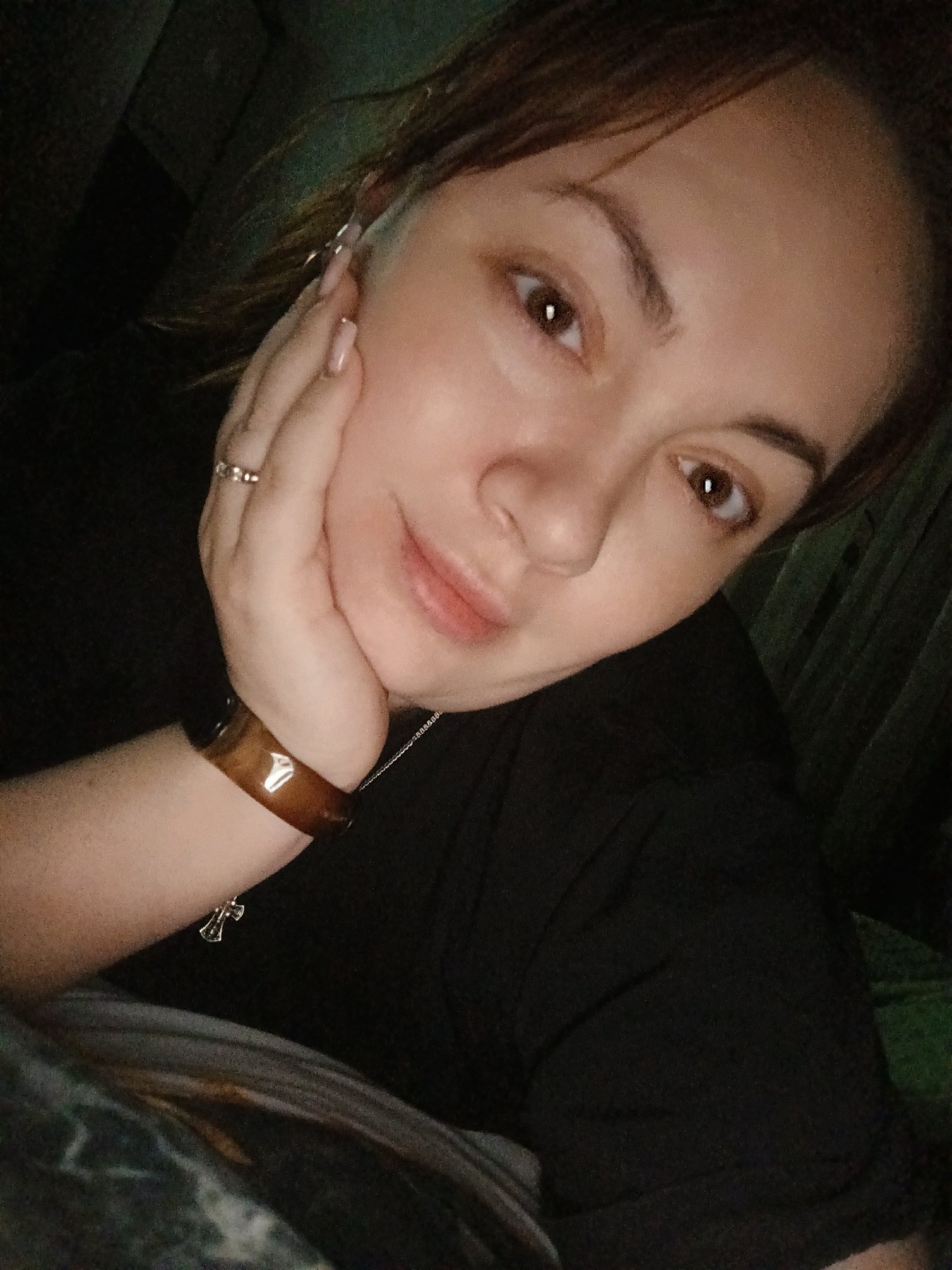 A medium close-up shot shows a woman with light skin, brown eyes, and brown hair. Her left hand is positioned on her cheek, with a silver ring visible on her ring finger. She is wearing a brown bracelet on her left wrist and a black t-shirt. A silver necklace is also visible. The background is dark, suggesting an indoor environment. The location is not identifiable as Chaikovsky, Russia based on the provided image.