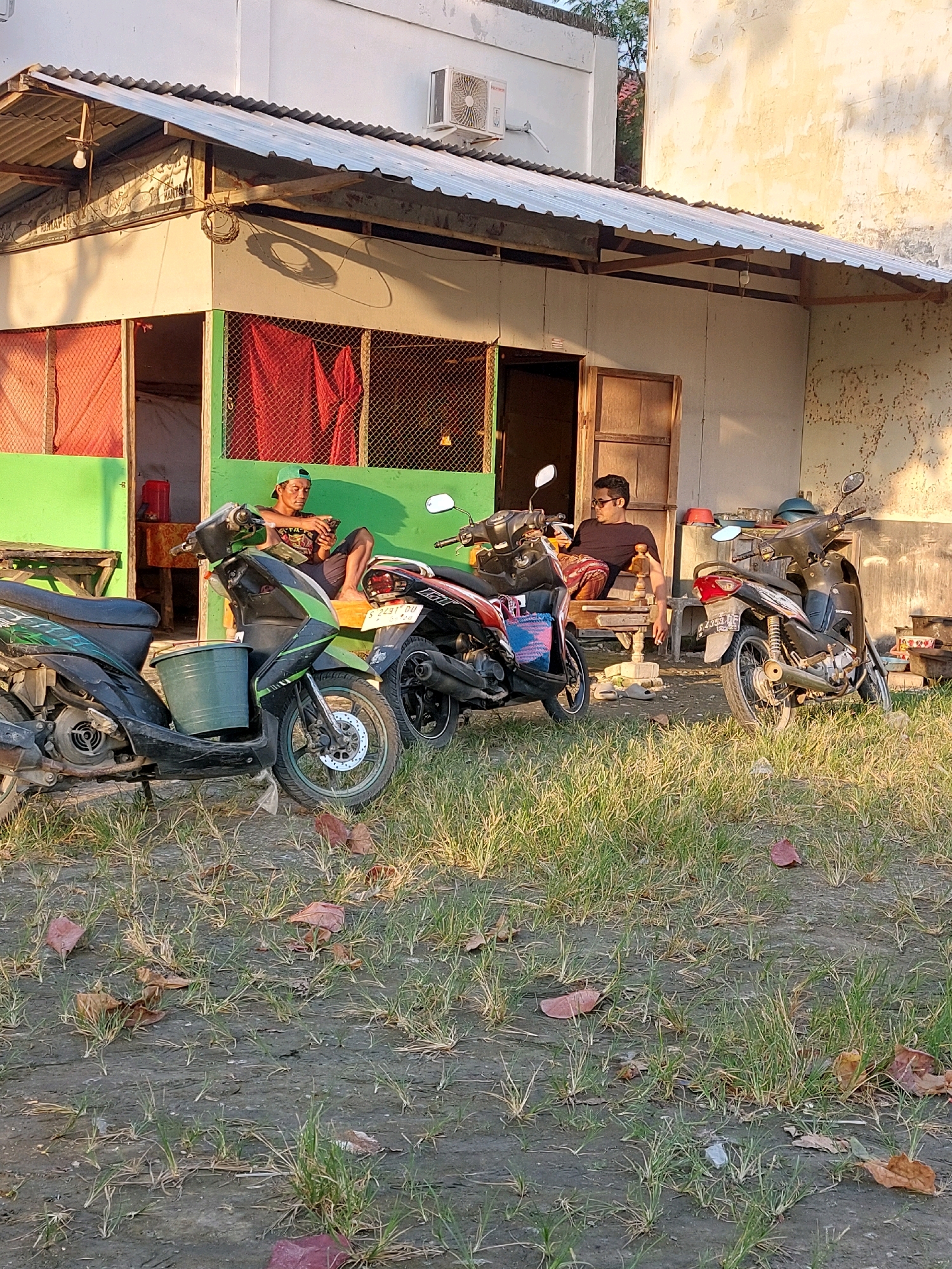 In SARANGAN, Indonesia, in front of a dilapidated, light-colored building with a corrugated metal roof supported by rusting metal beams, two men are seated on a bench outside. The building has green-painted trim and red fabric visible through a mesh-covered window on the left. An air conditioning unit is mounted high on the building's exterior wall.
The man on the left, wearing a green baseball cap, a dark t-shirt, and dark shorts, is looking down at his mobile phone. Beside him is a green bucket. Two motorcycles are parked in front of the building. The motorcycle closest to the viewer is black and green, with a green bucket attached to its side. Another motorcycle, dark in color, is parked to the right of the first.
The man on the right, wearing sunglasses, a dark t-shirt, and patterned pants, is also looking at his phone while seated on a wooden bench. A third motorcycle, dark in color, is parked to his right. The ground in front of the building is a mix of dry grass, dirt, and scattered brown leaves. The scene is illuminated by warm, late afternoon sunlight, casting shadows on the ground and the building.