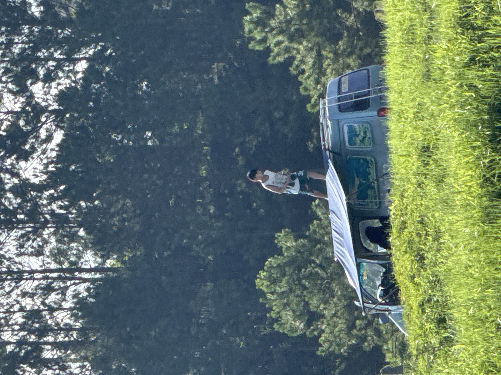 The image shows an outdoor scene in Da Lat, Vietnam, featuring a light blue or grey van partially obscured by bright green grass in the foreground. A young male, appearing to be a pre-teen or teenager, stands barefoot on the roof of the van. He wears a white sleeveless top and dark shorts, and holds an unidentified small object in his hands. A blue tarp is extended from the left side of the van, functioning as an awning, and a metal ladder is affixed to the rear of the vehicle, providing roof access. The van's front cabin is partially visible, with dark interior elements. In the midground and background, various pine trees are visible, with smaller, lighter green trees closer to the van and a dense, dark green forest dominating the distant horizon. Patches of bright white sky are discernible through the upper tree canopy.