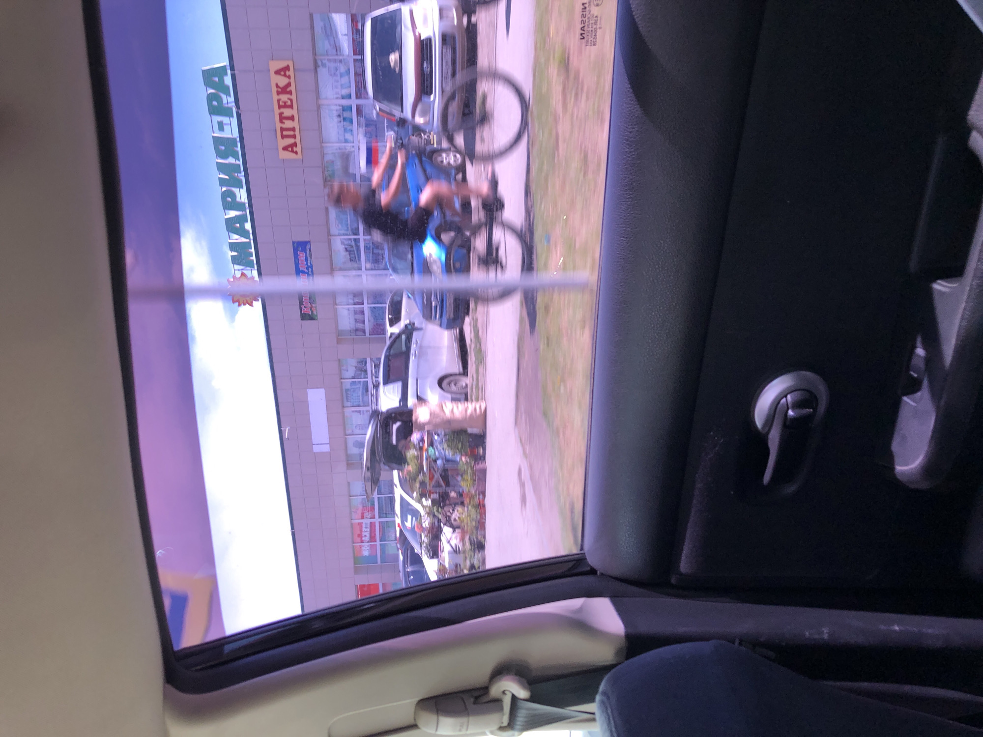 The image, taken from inside a vehicle, shows a street scene with a commercial building in the background. The building has signs that read "МАРИЯ-РА" and "АПТЕКА". A person is riding a bicycle in the middle of the street, blurred due to motion. Several cars are parked in front of the building and on the street. In the foreground, the interior of the car, including the door panel, seat, and seatbelt, is visible. The sky above is blue with some clouds. The overall lighting suggests it is daytime.
