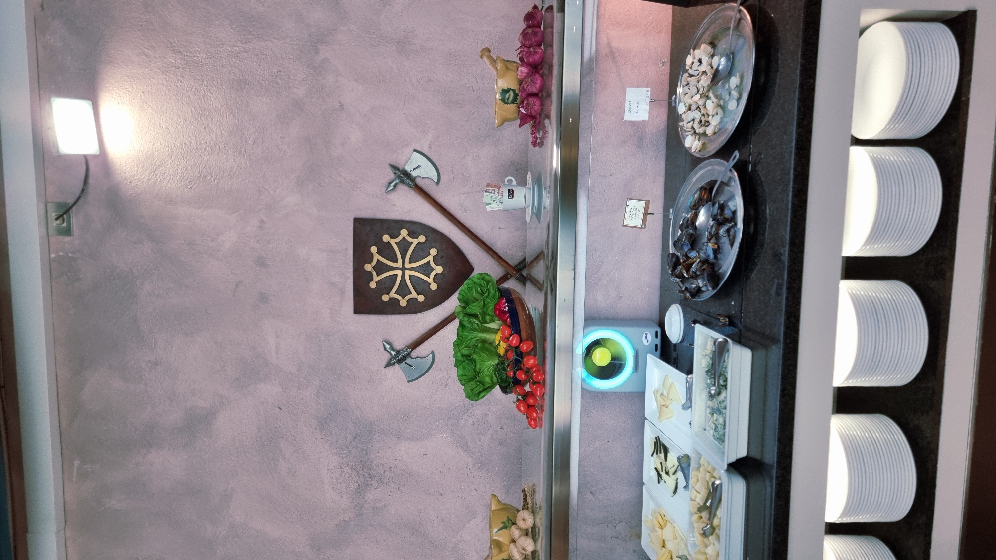 The image displays a buffet setup in what appears to be a restaurant or catering establishment. The background is a textured pink wall, with a spotlight illuminating the upper section. Centered on the wall is a shield-like emblem featuring a cross with four arms, flanked by two crossed axes.
On a shelf above the buffet counter, there are several items: a bowl of lettuce and tomatoes, a mortar and pestle with garlic and onions, and a cup of coffee with sugar packets.
Below this shelf, the buffet counter features various food items. To the left, there are trays of sliced cheeses. In the center, there is a tray of what appears to be a green dip or spread, next to a circular blue fan. Further to the right, there are two platters of seafood: one with mussels and another with clams. There are also small signs with handwritten text near the food.
Beneath the buffet counter, stacks of white plates are visible. The overall impression is of a well-organized food service area, possibly part of a larger establishment.
Regarding the specific location in Besalú, Spain, the provided image and its crops do not contain any explicit or implicit visual cues that would allow for its precise identification within the city. Therefore, the location cannot be determined from the image alone.