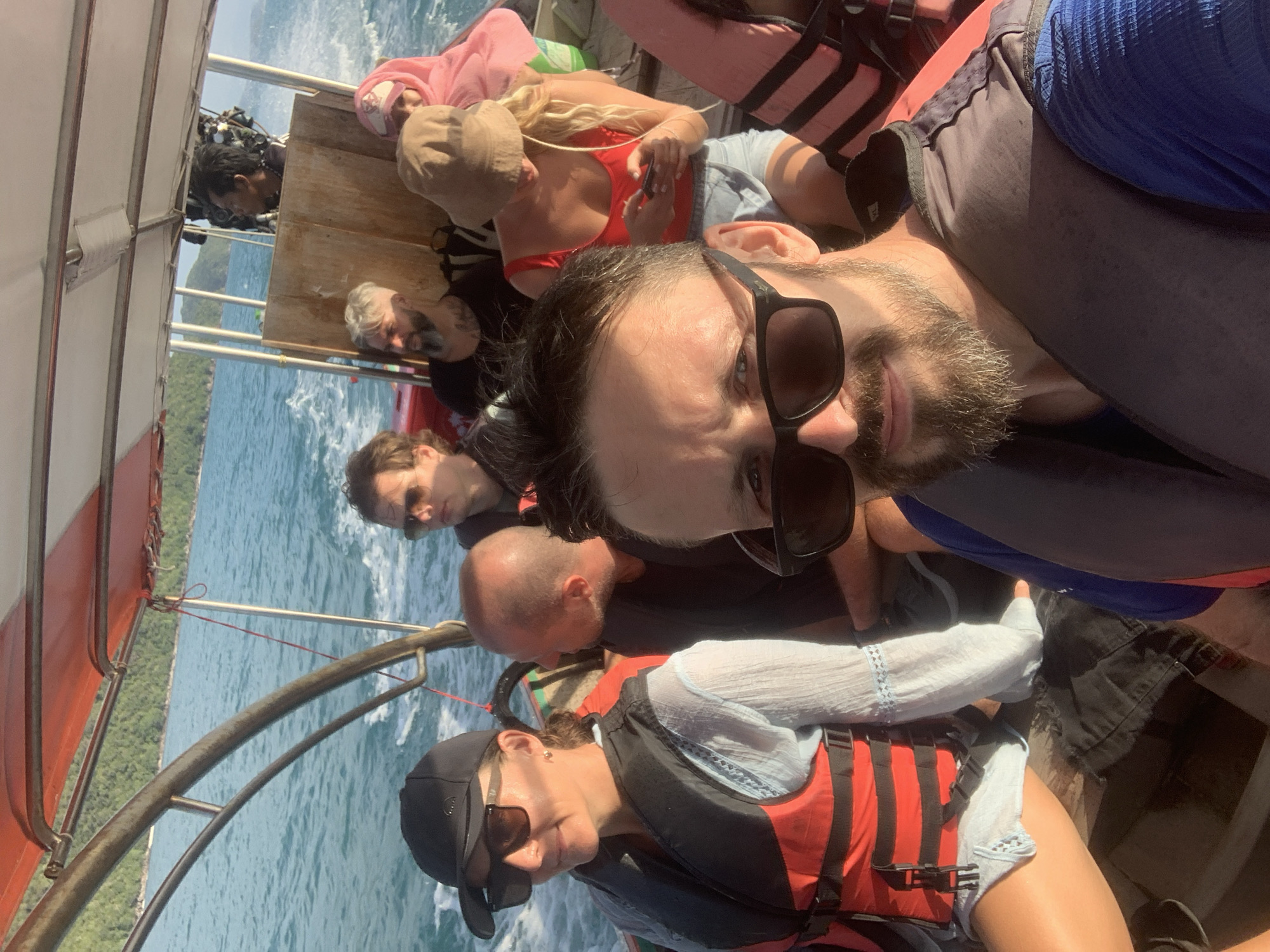 This is a medium-shot, front-facing selfie taken on a boat on a sunny day in the Ko Samui District, Thailand. The foreground is dominated by the face of a man wearing sunglasses and a dark life jacket. Behind him, multiple people are visible, also wearing life jackets, seated on the boat. The boat is traveling on a body of water with a green, tree-covered island visible in the background. The water is a turquoise blue, and sunlight is reflecting off its surface. The boat appears to be a small passenger vessel with a canopy overhead. The people are dressed in casual summer clothing, and some are looking towards the camera, while others are looking away. The overall atmosphere suggests a leisurely boat trip.