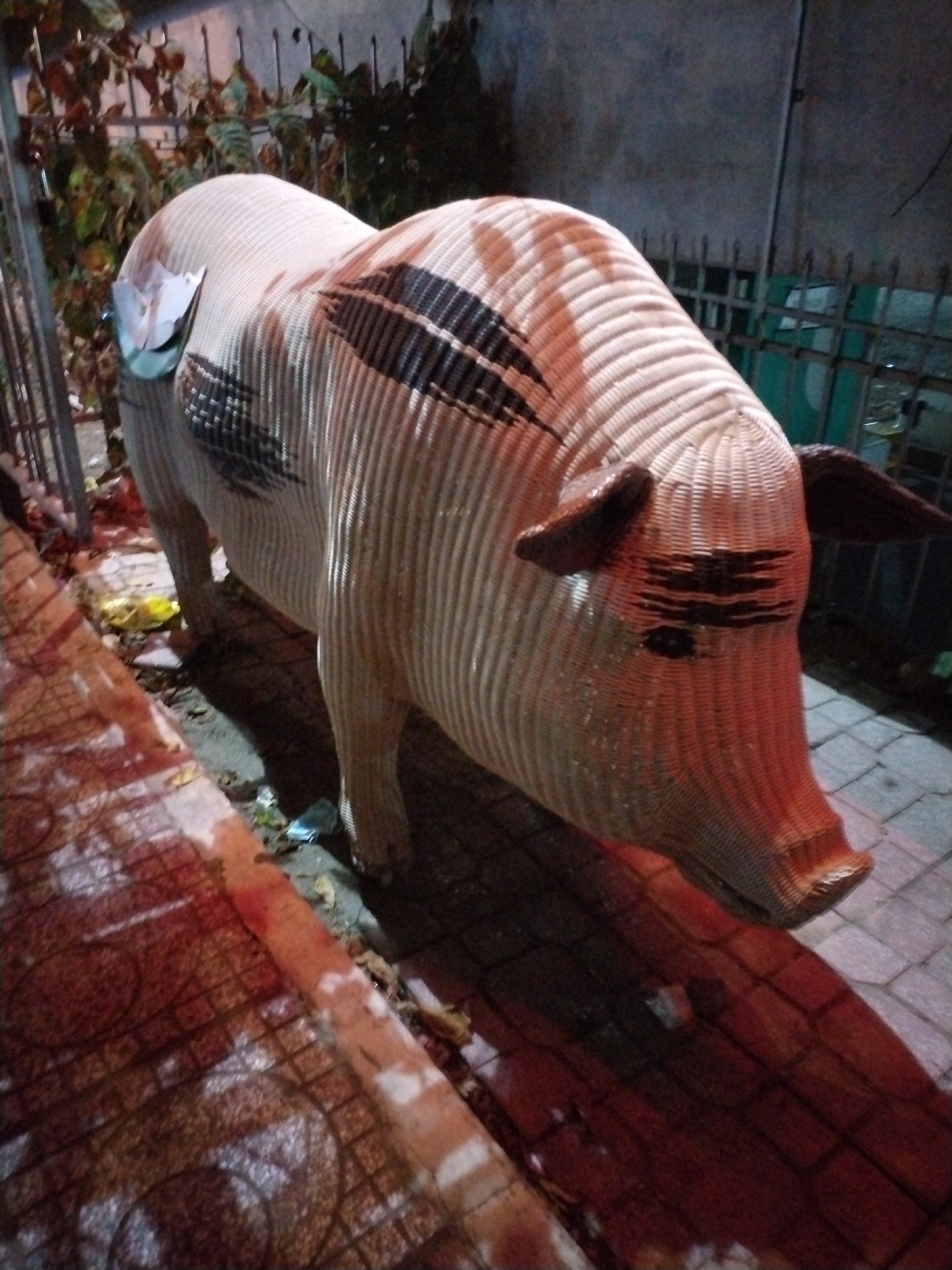 A wicker pig statue stands on a sidewalk in Bac Nha Trang Ward, Vietnam. The statue is primarily light beige with brown ears and snout, and dark brown markings across its body and face. It's positioned near a metal fence and some foliage. The sidewalk is paved with patterned tiles, and there's some debris and possible litter around the base of the statue. The lighting suggests it is either dusk or nighttime.