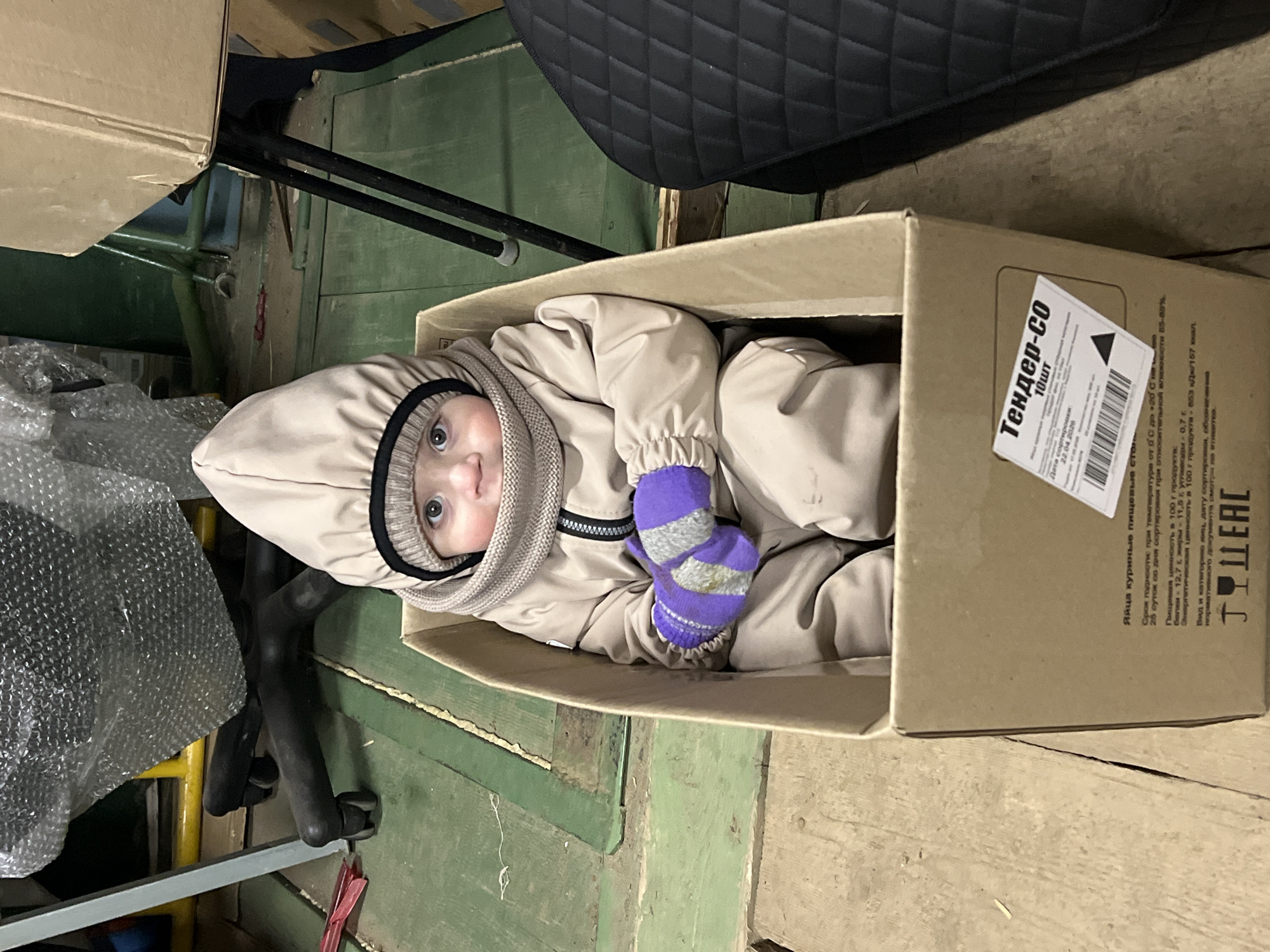 A baby in a beige winter suit and purple-and-gray striped mittens is sitting in a cardboard box on a worn, greenish-brown painted wooden floor. The box has a white label with Russian text, indicating "Tender-CO" and mentioning "chicken eggs" with a sorting date of "22.04.2026." The background includes a black padded seat, part of a chair frame, and packing materials.