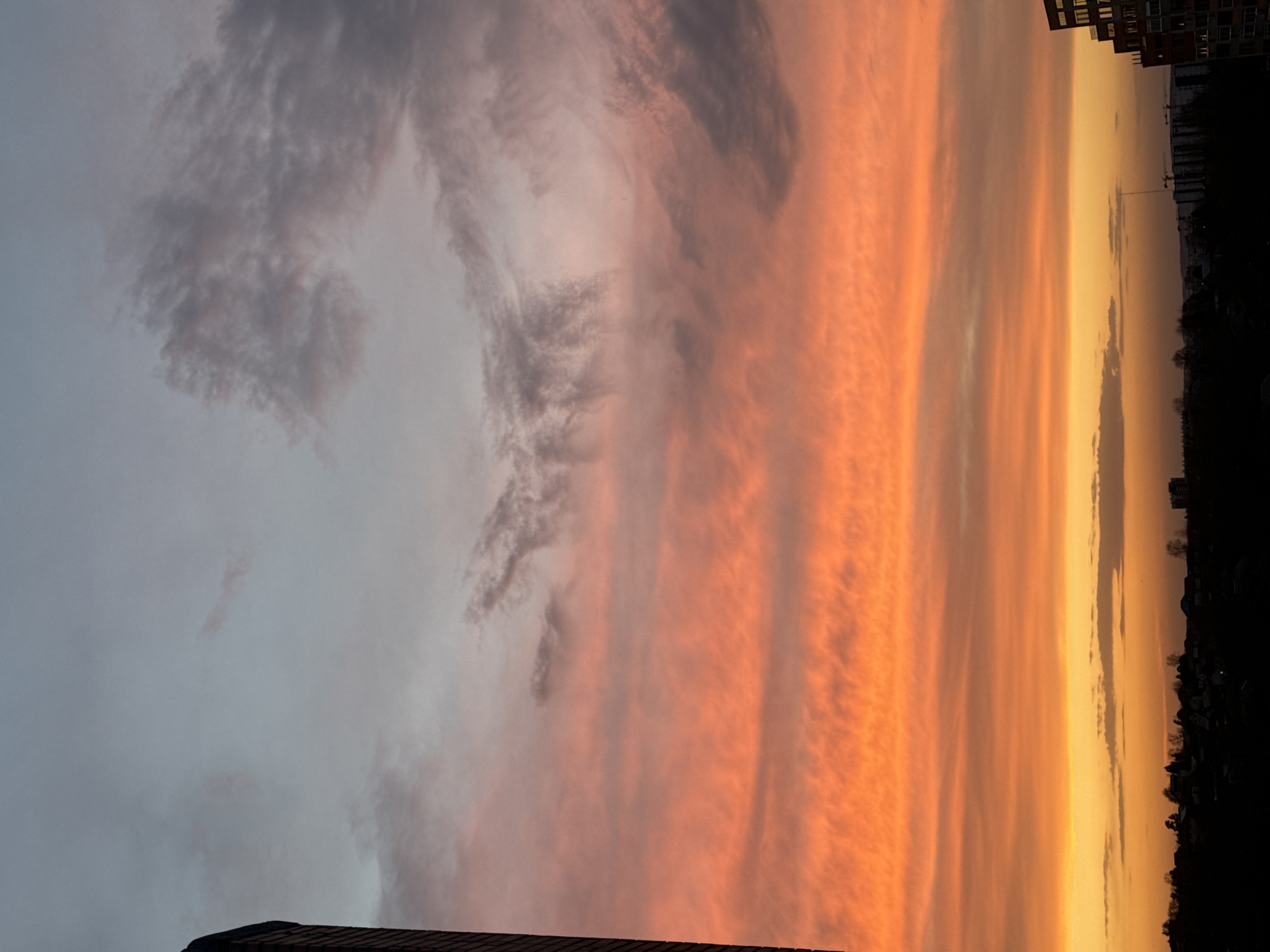 The image depicts a vivid sunset over Krasnoyarsk, Russia, with a gradient of colors transitioning from a cool gray in the upper left to warm oranges and yellows on the right. Wispy, textured clouds fill the sky, and in the bottom corners are the edges of buildings. The horizon is marked by silhouetted trees and the faint outlines of buildings, including apartment blocks. A tower crane is also visible against the lighter sky. The scene is urban, viewed from an elevated position.