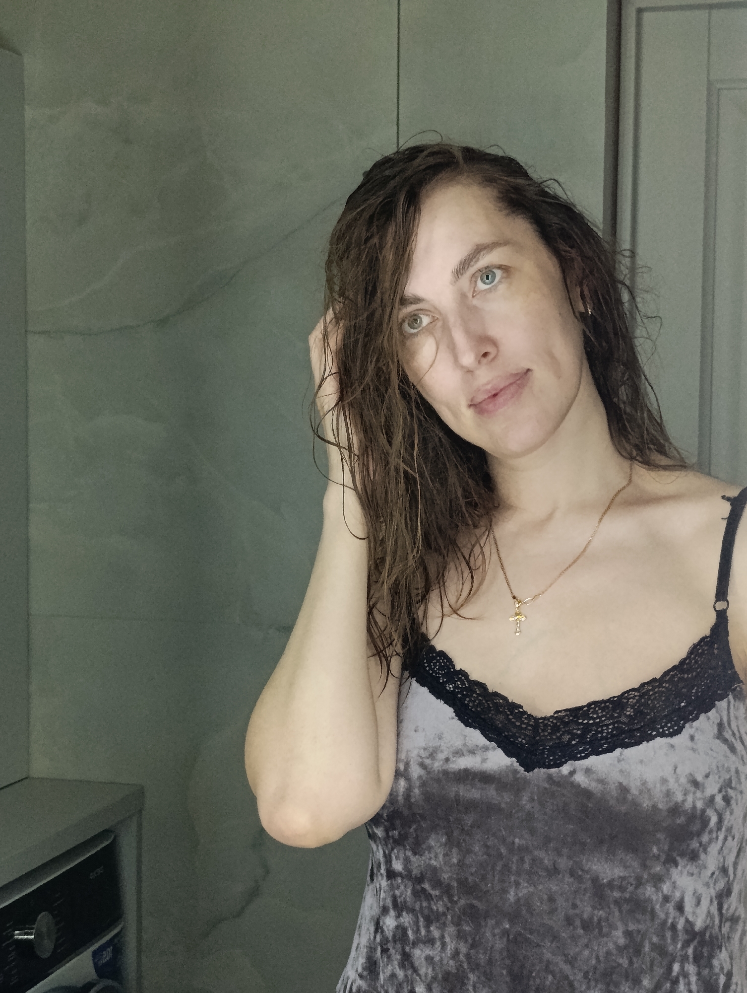 The image portrays a young adult woman indoors, likely in a bathroom or utility space, against a muted green-grey textured wall. Her medium-length brown hair appears damp or wet. She has fair skin and light-colored eyes, with one eye appearing blue/grey and the other green/hazel, suggesting heterochromia.
The woman looks slightly to her left (viewer's right) with a gentle, soft smile. Her left hand is raised, with fingers lightly touching her hair near her temple, as if casually pushing it back. She is wearing a greyish-purple crushed velvet camisole featuring black lace trim along the neckline. A thin gold chain necklace with a small, ornate gold cross pendant adorns her neck.
In the bottom-left corner of the background, a portion of a modern front-loading appliance, resembling a washing machine or dryer, is visible. The right side of the frame shows the edge of a door or cabinet. The lighting is soft and diffused, contributing to a calm and natural atmosphere. The scene suggests a domestic moment, possibly in Chaikovsky, Russia. There is no clearly legible text visible in the image.