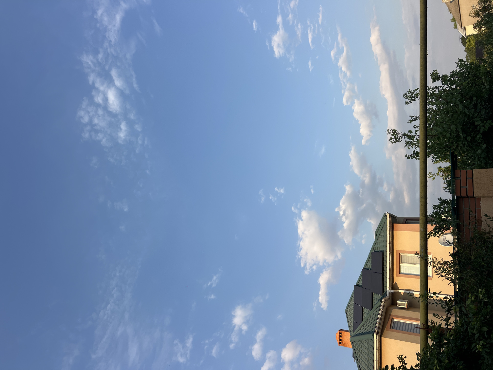 The image depicts a residential building with a green roof and solar panels, situated in the city of Fontanka, Ukraine. The building features a prominent brick chimney and several windows. In the foreground, a horizontal pipe runs across the frame, with lush green foliage visible below it, suggesting a garden or yard area. The sky is clear blue with scattered white clouds, indicating fair weather. No people are visible in the scene. The overall impression is a serene, daytime view of a suburban dwelling.