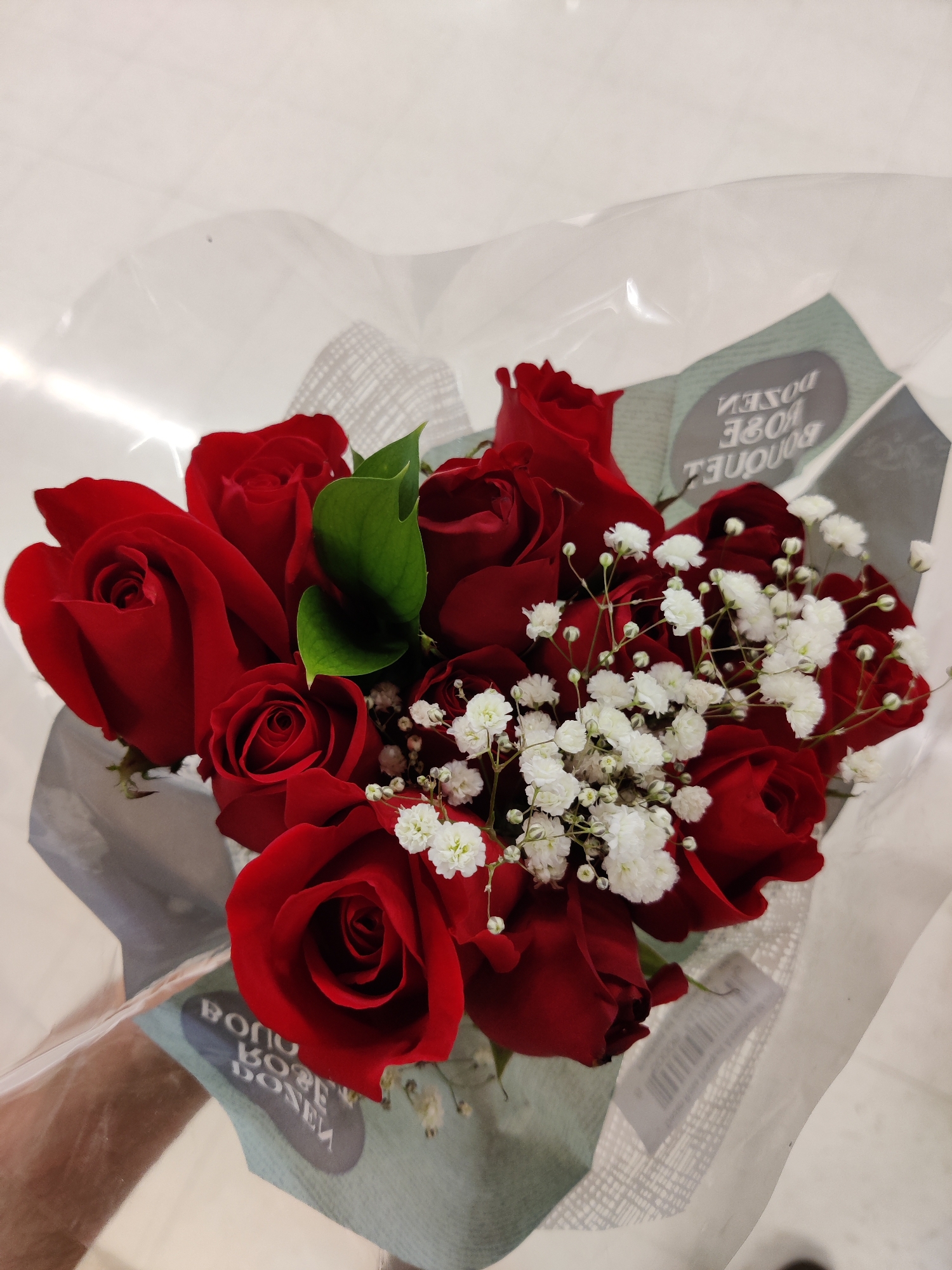 A close-up, overhead shot shows a bouquet of red roses with white baby's breath, wrapped in clear plastic. The bouquet is placed on a reflective surface that appears to be a floor, possibly in a store. Two oval tags with text are visible, one partially obscured by the flowers and plastic. The text on the visible tag is in reverse, reading "BOUQUET ROSE DOZEN." The lighting suggests an indoor environment, possibly a grocery store or flower shop. The image was captured in Mechanicsburg, United States of America.