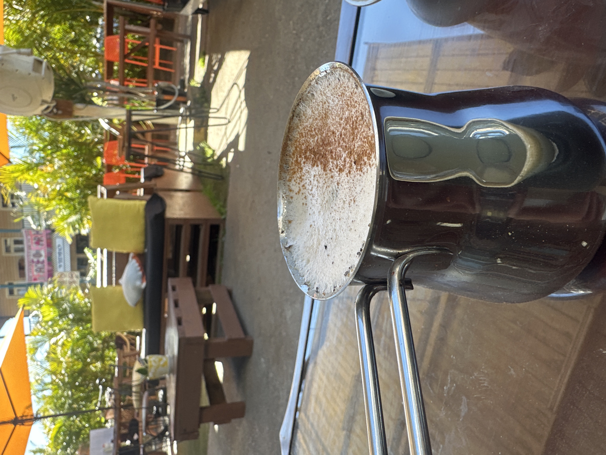 The image shows a close-up of a coffee pot with frothy coffee topped with cinnamon, resting on a wooden surface. In the background, an outdoor seating area is visible, with tables, chairs, and lush green foliage under an orange umbrella. The scene appears to be an outdoor cafe. The presence of banners with text that appear to be in English, such as "ACE OF ARC" and "VEGAS", along with the style of the outdoor seating and vegetation, suggests a casual, possibly tropical or subtropical setting. The general atmosphere is relaxed and sunny. Given the question about the location in Bellingen, Australia, this image likely depicts a cafe or restaurant in that area, known for its natural beauty and outdoor dining options.