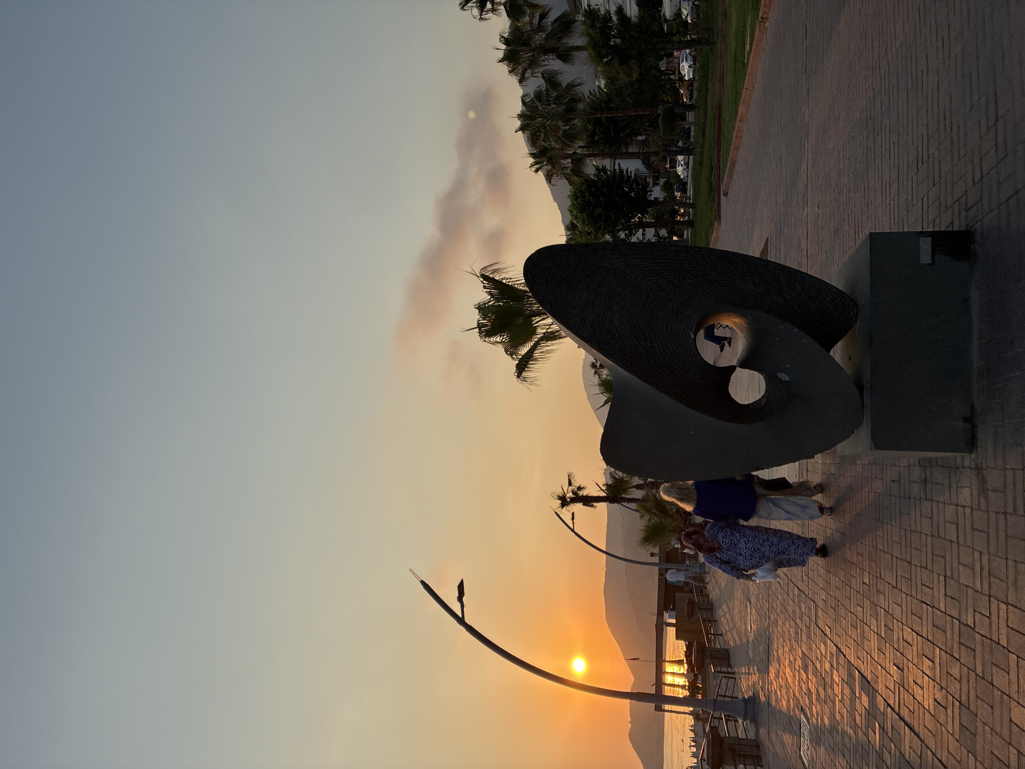 This is a photograph taken at dusk in Alanya, Turkey, likely along the waterfront promenade. The setting sun casts a warm, orange glow across the scene, illuminating the sky and reflecting on the water. In the foreground, a large, abstract dark sculpture with a circular opening is prominent. Two women are walking away from the camera on a paved walkway made of brick-like stones. One woman wears a blue and white patterned dress, and the other wears a blue top and light-colored pants. Behind them, palm trees are visible, along with a street lamp and a portion of the sea. In the distance, hills are silhouetted against the sunset. The overall atmosphere is peaceful and serene, indicative of an evening stroll in a coastal city.
