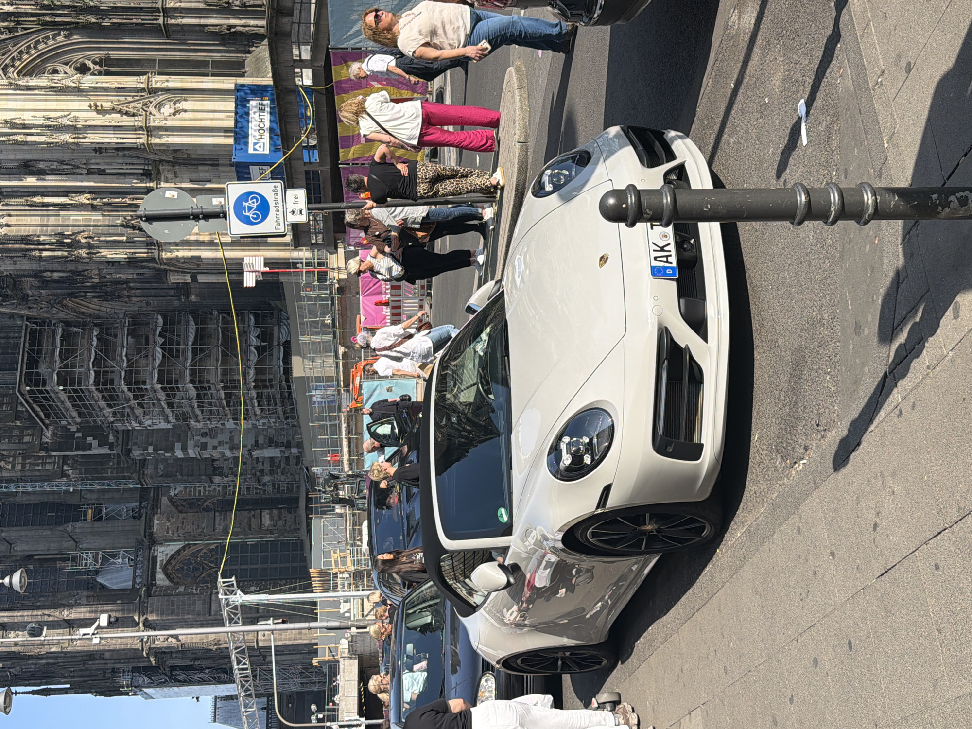 The photograph captures a street scene in Cologne, Germany, with the iconic Cologne Cathedral prominently featured in the background, currently undergoing construction or renovation as scaffolding is visible. A white Porsche is parked along the street, drawing attention due to its sleek design. Pedestrians are present, some walking, others appearing to be tourists observing the cathedral, showcasing a mix of local and visitor activity. A bike lane sign is present.
