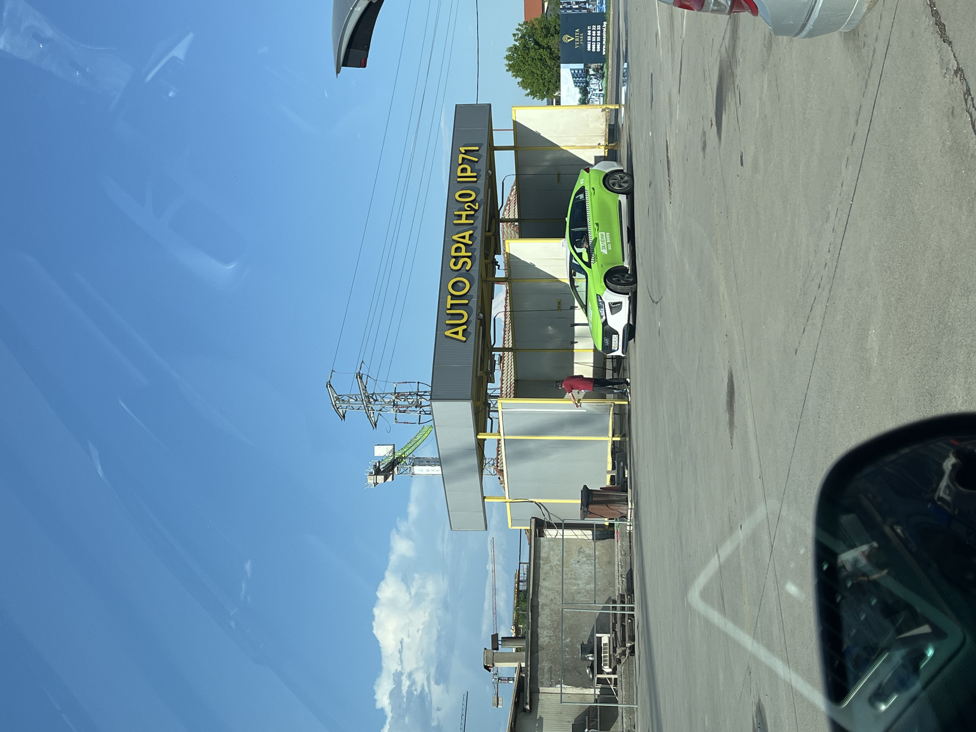 This image depicts an outdoor car wash facility named "AUTO SPA H2O IP71" in Plovdiv, Bulgaria, specifically at ул. "Васил Левски" 222, 4003 Northern Industrial Zone. The scene is observed during daytime under a predominantly clear blue sky with scattered white clouds.
In the center, a bright green and white Hyundai Ioniq, identifiable as a taxi by the "ЗЕЛЕНИ" (Green) text and phone number "002 9999" on its side, is positioned within one of the car wash bays. A Bulgarian license plate, partially visible as "PB 6245 KC", is affixed to the vehicle. A single male individual, wearing a red short-sleeved shirt, dark pants, and a dark cap, stands near the front driver's side of the green taxi, holding a spray nozzle and actively washing the car.
The car wash structure features light gray corrugated metal panels with yellow support frames and an overhead sign displaying "AUTO SPA H2O IP71" in yellow letters. To the left of the car wash, an industrial or construction area is visible, including concrete structures with a chimney, metal fencing, and multiple construction cranes in the distance. To the right, a large billboard advertises "VERITA HOME" by "MAXISTROI," displaying building renderings and contact information, with trees beyond it. Overhead, power lines and a large metal electricity pylon are visible, along with another construction crane with a prominent green jib. The ground consists of asphalt pavement with some visible stains. The image is taken from inside a vehicle, with reflections visible in the upper sky and a black side-view mirror capturing interior elements in the lower-left foreground. The rear bumper of a silver vehicle is partially visible in the lower-right foreground.