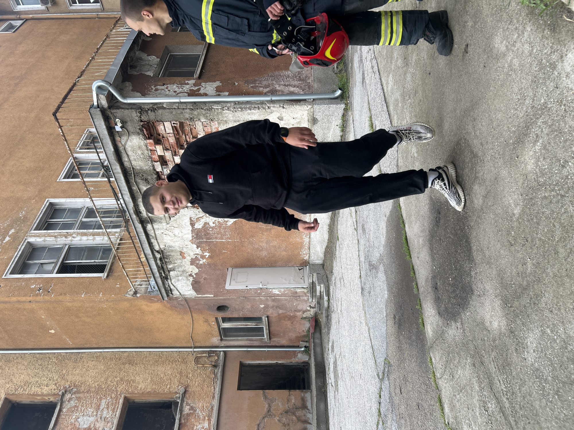 This image depicts two men in an urban alleyway or passage between old buildings in Sofia, Bulgaria, during what appears to be an overcast daytime.
In the foreground, a young man with short hair is posing for the camera, looking directly at the viewer with a neutral expression. He is dressed in a black hoodie, which features a small logo on the front (likely "TOMMY HILFIGER"), and black sweatpants. He is wearing distinctive white and black striped sneakers, with the text "SPLY-350" visible on the side stripe.
Behind him, partially obscured, stands another man dressed in what resembles a firefighter's uniform, dark blue or black with yellow reflective stripes on the arms and legs. He is holding a red and yellow helmet in his hands. His face is not clearly visible, and he appears to be looking downwards or away from the camera.
The background consists of old, weathered buildings. The structure to the left has a faded, brownish facade with extensive peeling paint, revealing exposed brickwork underneath. Multiple windows, some partially open, are visible, along with a metallic drainage pipe running down the wall. The ground is a cracked concrete or asphalt surface, with small tufts of grass growing in the fissures. The lighting is soft and diffused, indicating an overcast sky and no strong shadows. The overall scene conveys a gritty, lived-in urban environment.