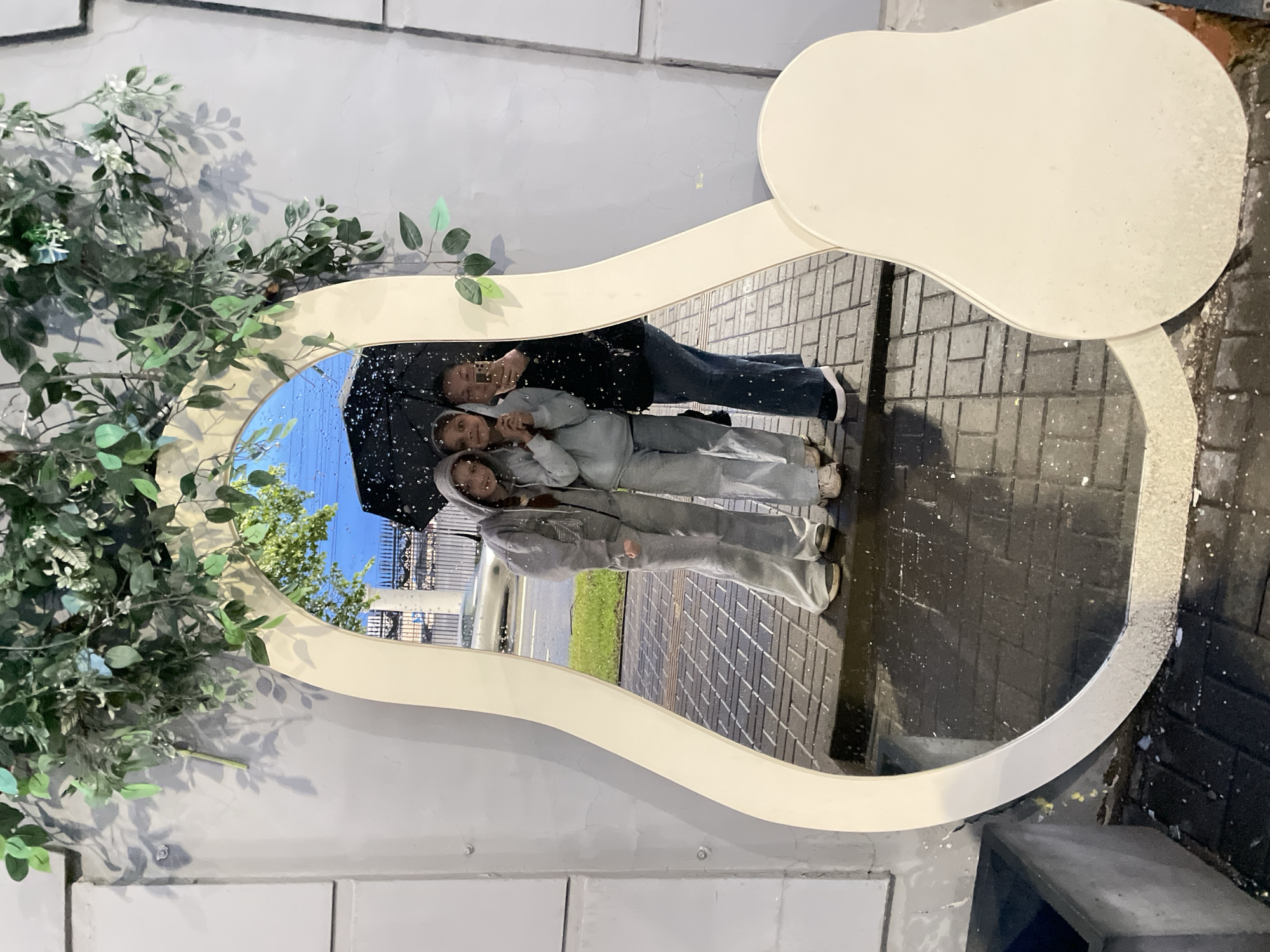 Here's a description of the image:
A pear-shaped mirror with a wide, white frame is mounted on a wall constructed of light gray, vertically-oriented tiles. To the left of the mirror, artificial foliage is attached, partially obscuring the frame. The reflection in the mirror shows three people standing outdoors under an umbrella, suggesting rainy weather. The background of the reflection includes a street, a sidewalk paved with gray bricks, and some greenery. The location is Izhevsk, Russia.
