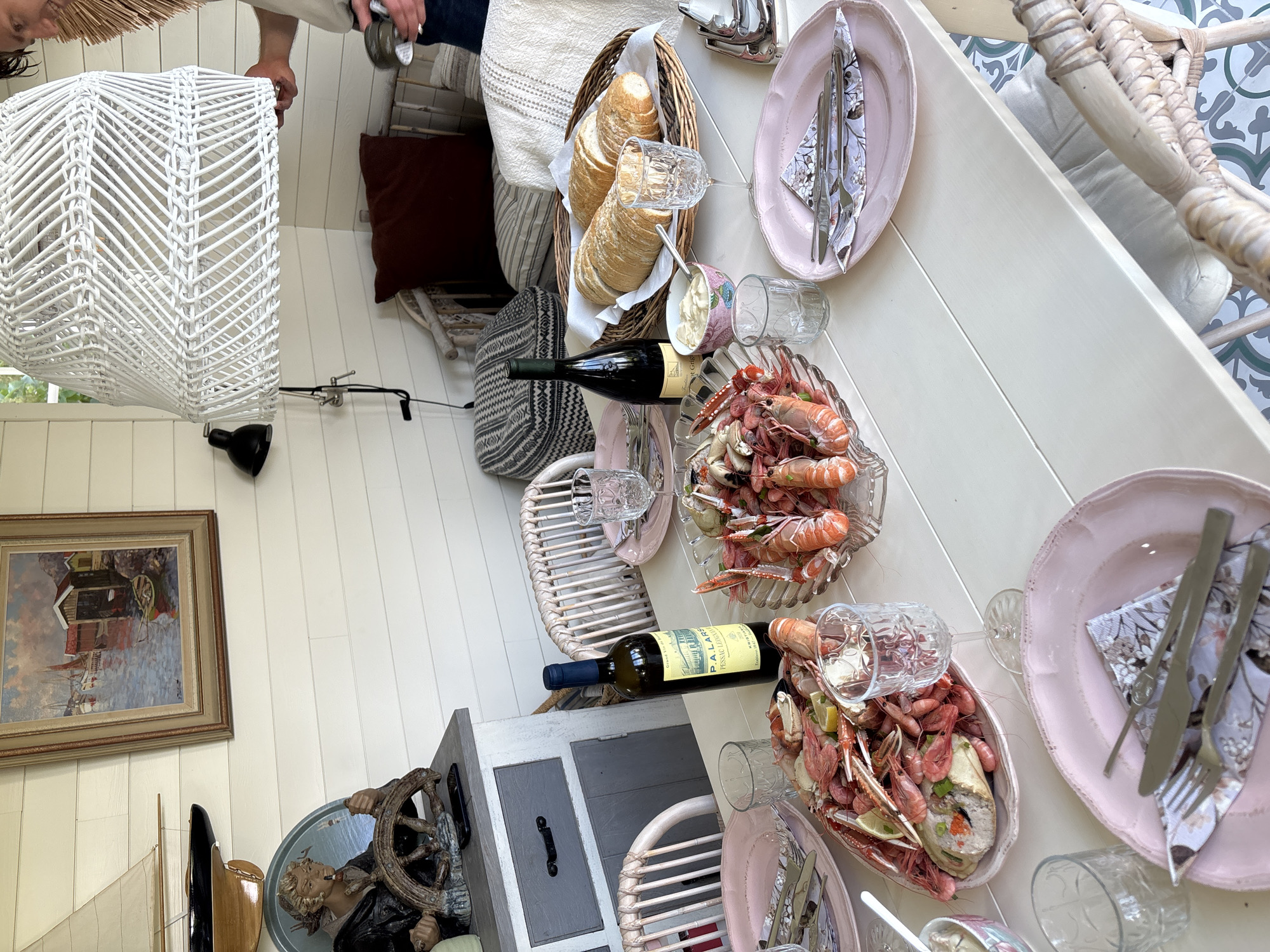 The image depicts an outdoor dining setting on a table laden with seafood, bread, and drinks, likely a terrace or patio. A large white rattan lampshade hangs overhead, and a person's arm is visible on the right, holding a napkin and a silver goblet. The table is set with pink plates, cutlery, and patterned napkins. Several dishes of seafood, including shrimp and langoustines, are prominently displayed. There are also glasses and a bottle of wine. In the background, a painting of a harbor scene hangs on a white paneled wall, and decorative items such as a ship's wheel and a figurine are visible. Wicker chairs surround the table. The floor has a patterned tile design. Based on the provided location of Larkollen, Norway, the scene is situated in this coastal town known for its scenic beauty and maritime activities.