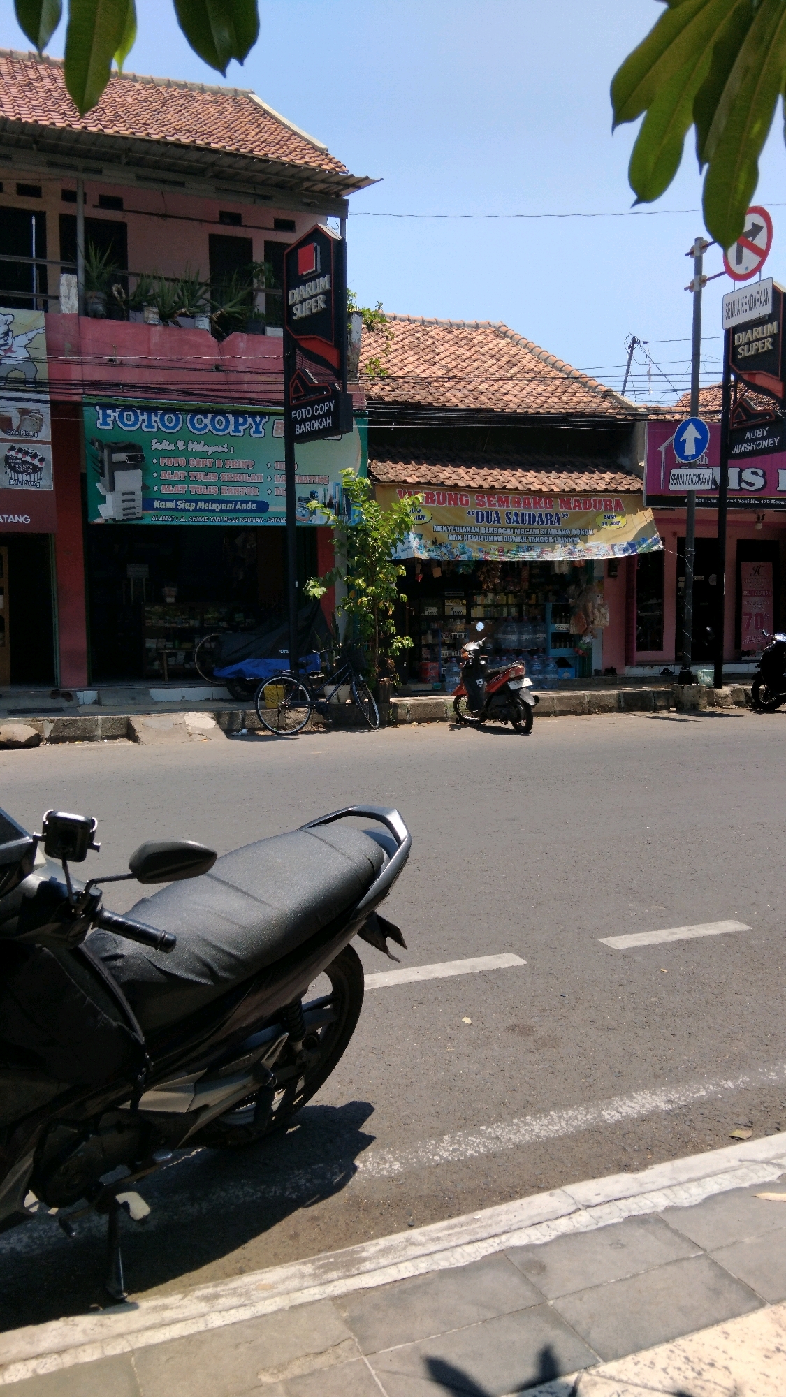 A street scene in Batang, Indonesia, is captured under a clear blue sky. The foreground features the rear of a parked black motorcycle. The middle ground depicts a street with a white painted line dividing the lanes, and a sidewalk. Beyond the sidewalk, buildings line the street. One building, on the left, is painted pink and has a "FOTO COPY BAROKAH" sign. Next to it, another building with a red tiled roof has a "DIARUM SUPER" sign. A banner for "WARUNG SEMBAKO MADURA 'DUA SAUDARA'" hangs above a store displaying various goods. Further down the street, a traffic sign for "DIARUM SUPER" and "SEMUA KENDARAAN" is visible. Several other motorcycles are parked or in motion in the background, and a bicycle is propped against a building. The environment appears to be a commercial area with shops and signs in Indonesian.