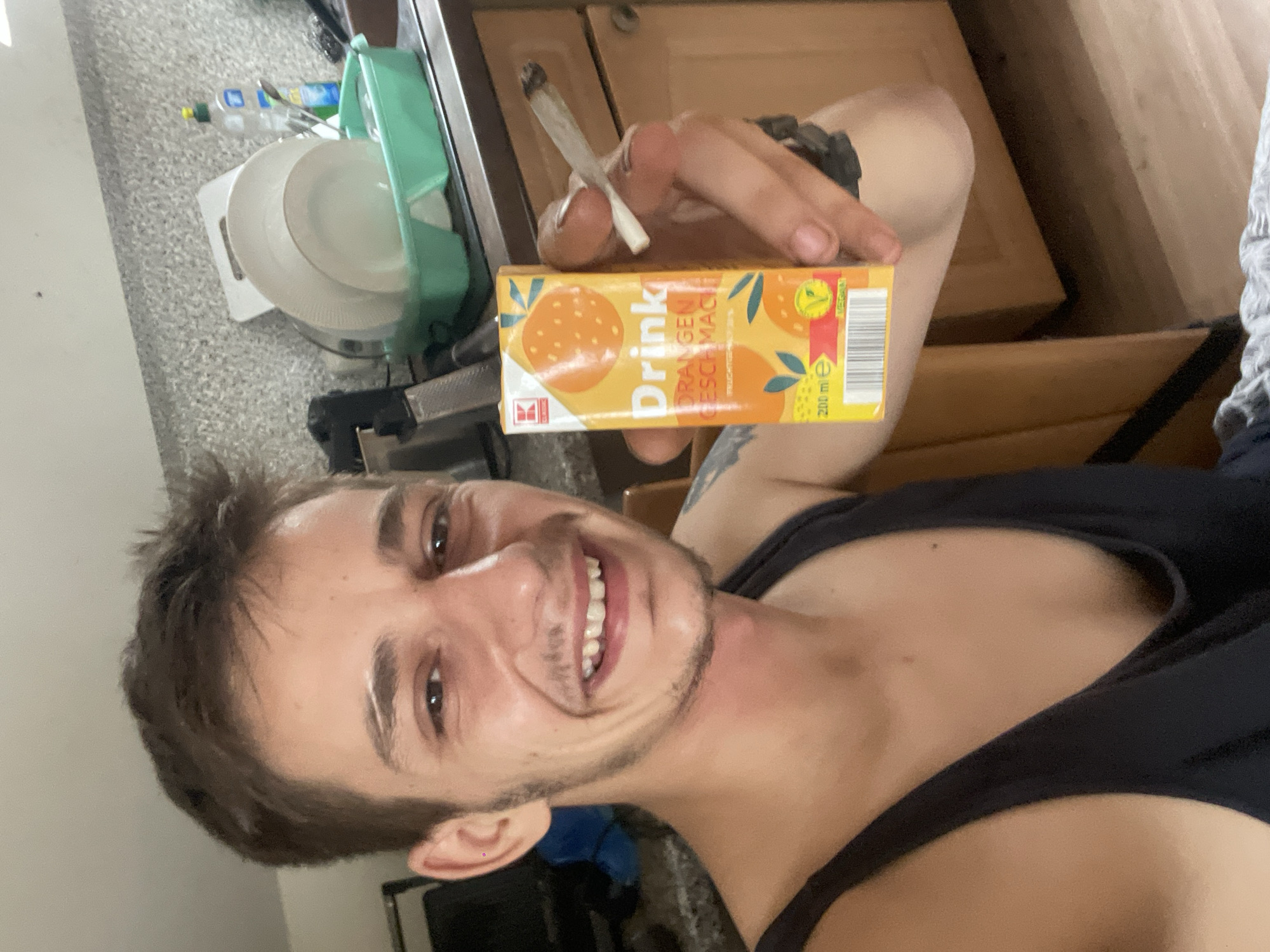 The image shows a close-up of a young man with short brown hair and a light beard, smiling at the camera. He is wearing a black tank top and has a tattoo on his left arm. In his right hand, he holds a small carton of orange juice and a lit cigarette. The background appears to be a kitchen, with wooden cabinets, a countertop, and a dish rack with plates and a bottle of dish soap. The lighting is moderate, and the focus is primarily on the man and the objects he is holding. The scene suggests a casual, indoor setting.