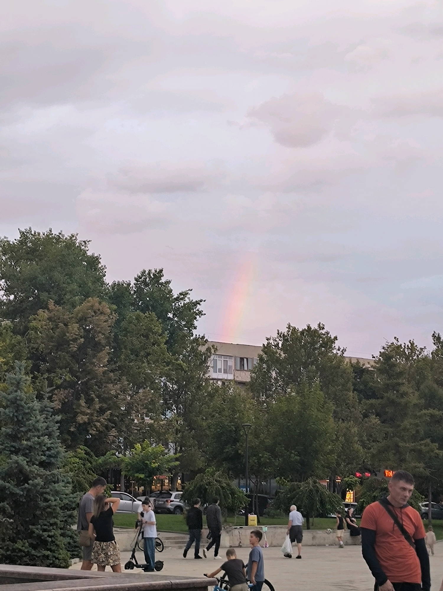 The image depicts an outdoor scene, likely in a city park or public square, with a subtle rainbow visible in the overcast sky. In the foreground, several people are present. A couple, a man and a woman, stand near a blue spruce tree. The woman is wearing a patterned skirt and a black t-shirt, while the man is in a grey t-shirt. Nearby, a young person on a scooter and another person with a bicycle are also visible. Further in the background, a group of people are walking, and one individual in a bright orange t-shirt is prominently positioned on the right side of the frame. Lush green trees form a backdrop, with a building visible behind them. The overall atmosphere suggests a casual, public gathering. The presence of the rainbow indicates a recent or ongoing rain shower. The location is identified as Chișinău, Moldova.