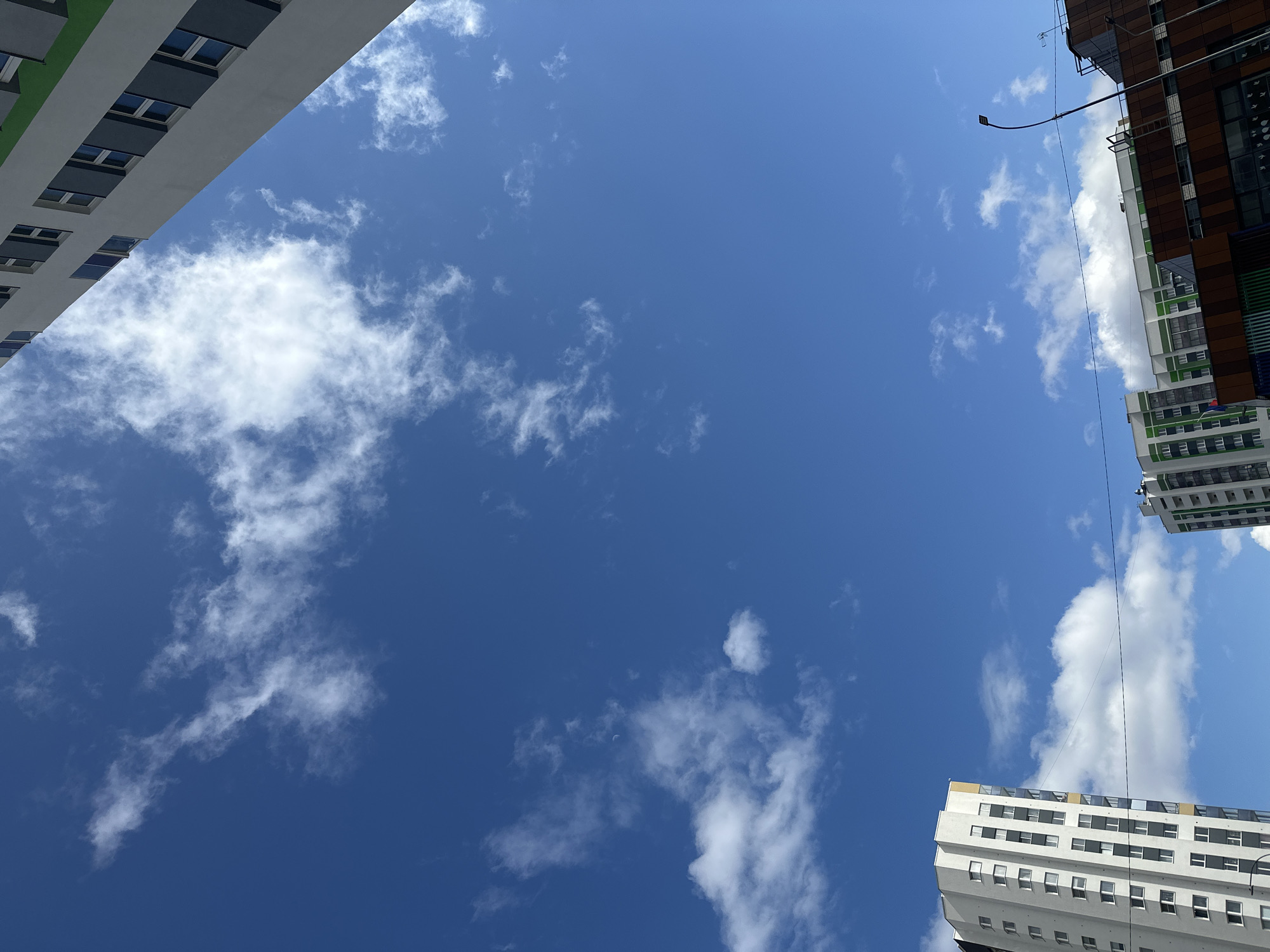 The image presents a worm's-eye view looking straight up at a vibrant blue sky, framed by sections of three modern high-rise buildings. The sky is largely clear, dotted with scattered, wispy white clouds of various sizes.
On the left, a portion of a building with a grey facade and numerous dark-framed windows is visible, featuring a touch of green on its top edge. In the upper right, part of a building with brown and dark-toned cladding, along with green and white elements, is seen. A curved street lamp extends into the frame near this building, accompanied by thin utility lines crisscrossing the sky. The bottom-right edge of the frame reveals the top of a white building, distinguished by rows of small windows, grey paneling, and a narrow yellow band along its roofline.
A notable detail in the expanse of blue sky is a faint, crescent moon, discernible slightly to the right of the image's center. The bright, even lighting indicates it is daytime, likely morning or afternoon. The overall scene conveys an urban setting in Yekaterinburg, Russia, focusing on the interplay between modern architecture and the open sky. There is no visible text or explicit human activity.