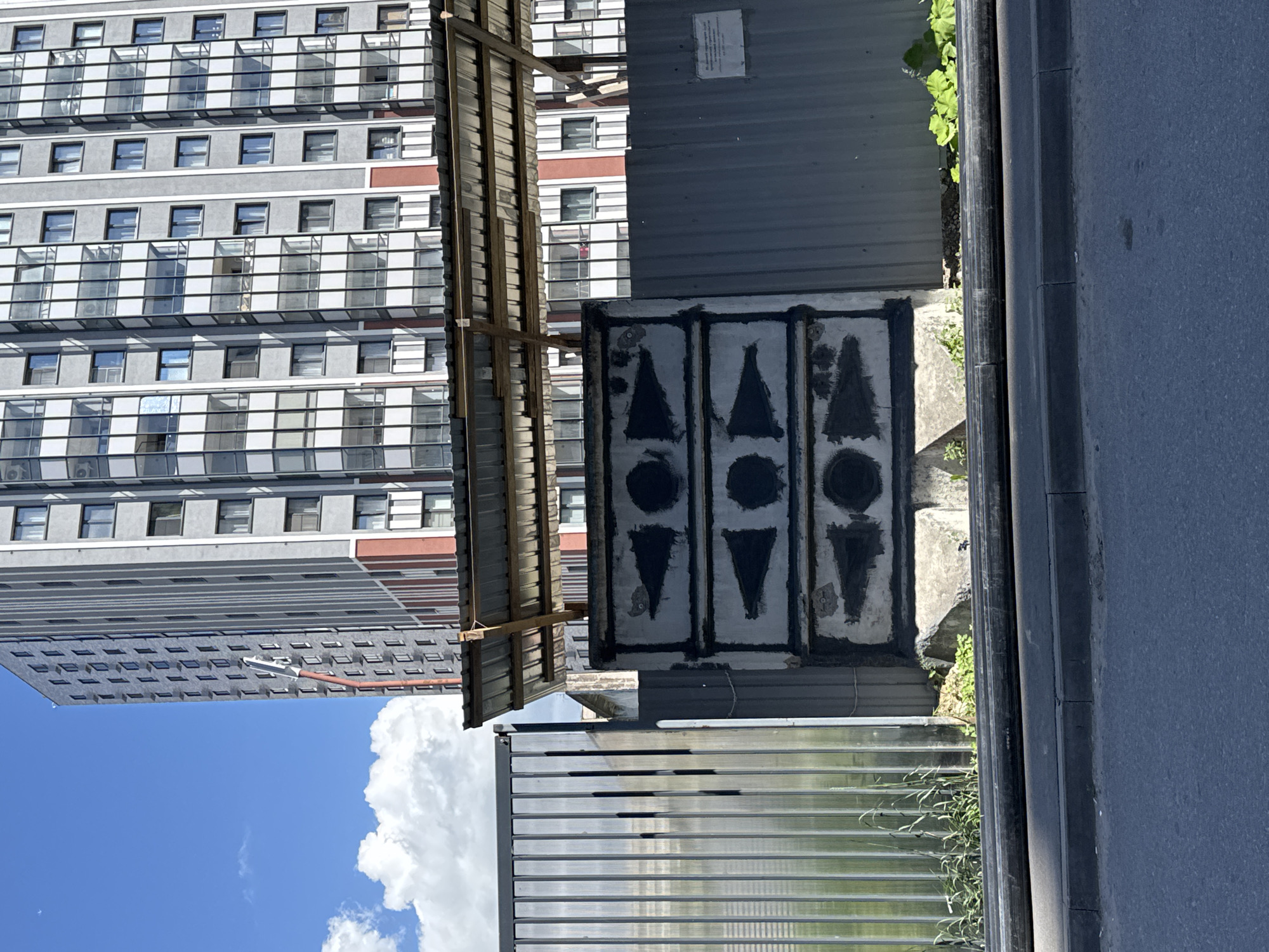 The image depicts an outdoor urban scene in Saint Petersburg, Russia, with a tall, modern residential building dominating the background under a partly cloudy blue sky. The building features a gray facade with vertical white and red stripes, and numerous windows and balconies. In the foreground, a dark asphalt road meets a gray concrete curb. A black pipe runs horizontally along the base of a metal fence. The fence itself is composed of vertical corrugated metal panels on the left and corrugated sheets on the right, with a partial corrugated metal roof supported by wooden beams extending over a concrete structure. This concrete structure has three rectangular sections, each containing two black arrow shapes pointing towards a central circle, with a solid black circle to the right of the arrows in the top and bottom sections, and a solid black circle to the left of the arrows in the middle section. Green vegetation grows around the base of the concrete structure and the fence.