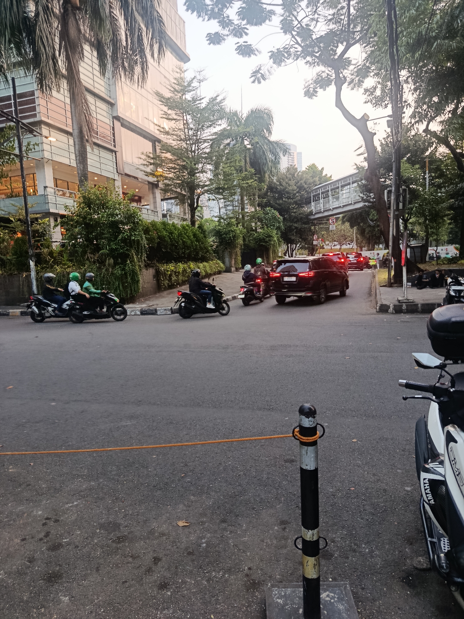 This image depicts a street scene in South Jakarta, Indonesia. In the foreground, a black and white bollard with orange rope attached is visible on the asphalt road. To the right, the front of a parked white and black motorcycle with "YAMAHA" branding is partially seen.
In the middle ground, several motorcycles and a black SUV are on the road. Three motorcycles are traveling side-by-side in the left lane, each with at least one rider wearing a helmet. One motorcycle further ahead is also occupied by a rider in a helmet. A black SUV is in the right lane, heading away from the viewer. In the background, a modern building with glass windows and balconies stands on the left, surrounded by lush green trees and palm trees. A pedestrian bridge with white railings spans over the road further in the distance. Traffic lights are visible. The sky is overcast, suggesting either early morning or late afternoon.