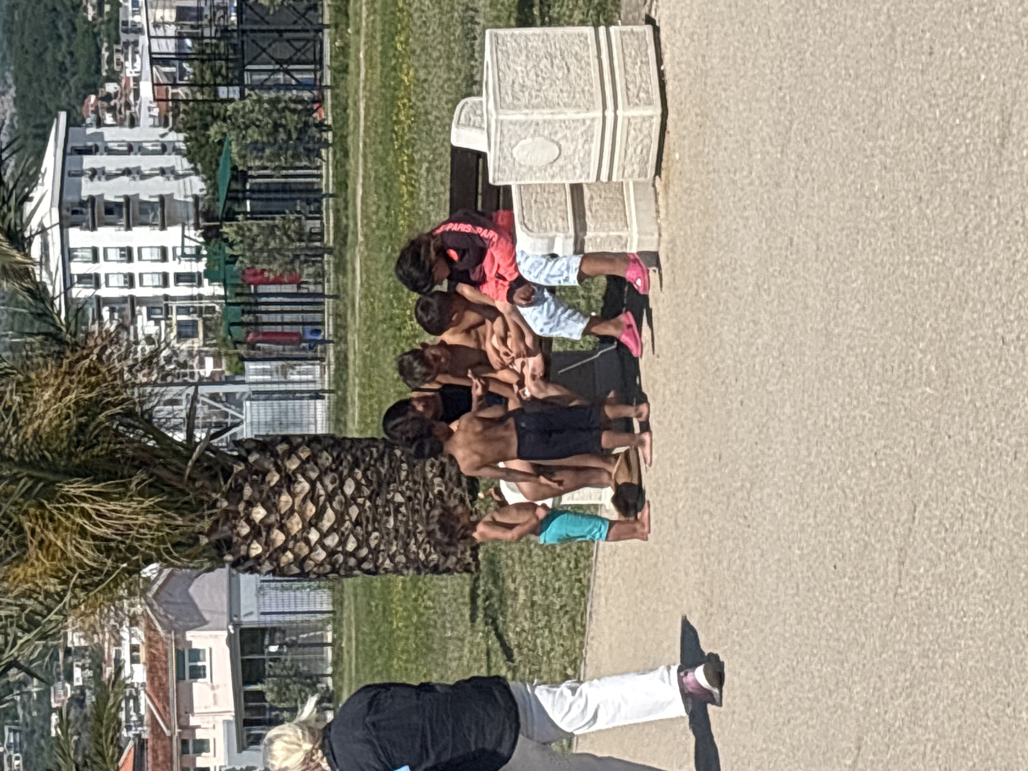 The image shows a group of children gathered by a bench. Some are sitting on the bench and others standing near it. A woman in a black shirt and white pants is walking past them on the sidewalk. A palm tree stands nearby. In the background, there are buildings and a playground. The location is in the city of Bar, Montenegro.